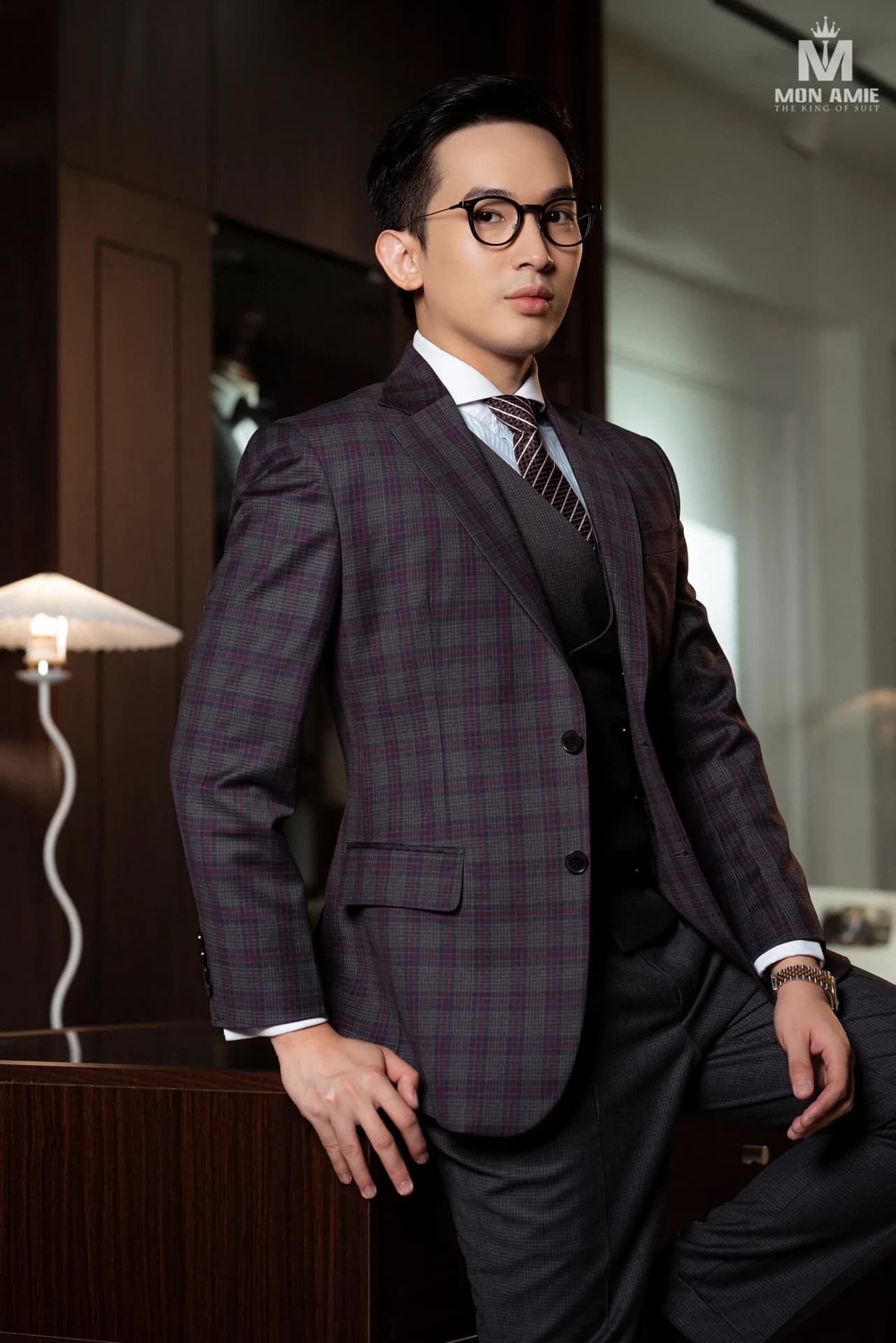 Suit ba mảnh caro tím xám, cà vạt sọc, dáng tựa bàn gỗ trong phòng ấm