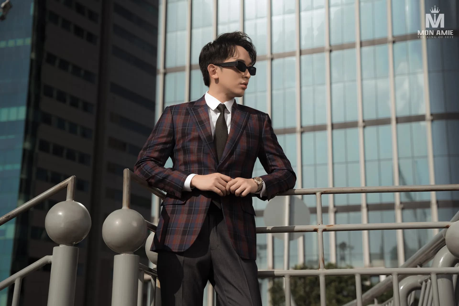 Blazer caro tím nâu phối quần xám, dáng tựa lan can giữa các tòa nhà kính