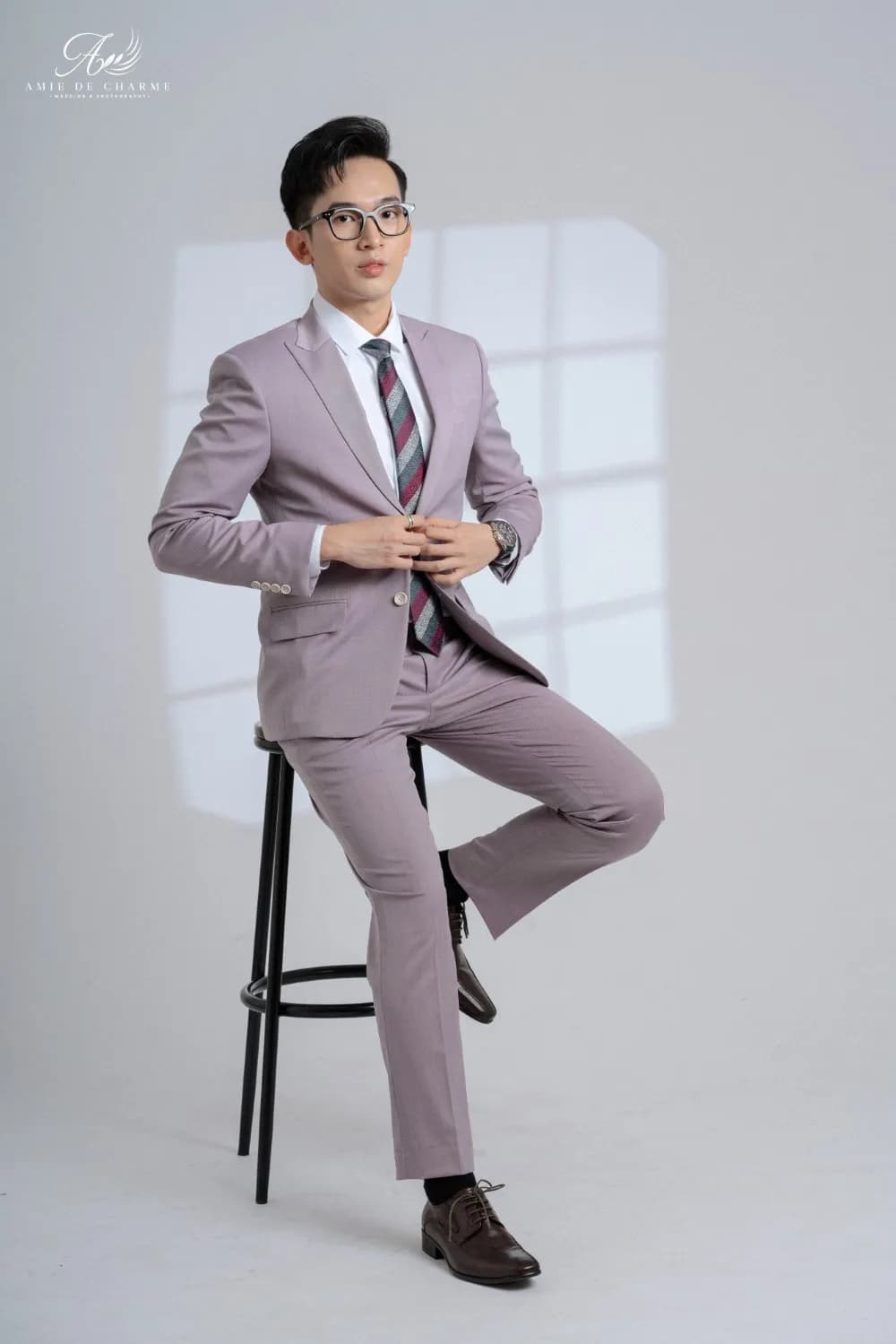 Suit tím hồng nhạt, cà vạt sọc, ngồi ghế cao chỉnh khuy trên nền studio xám