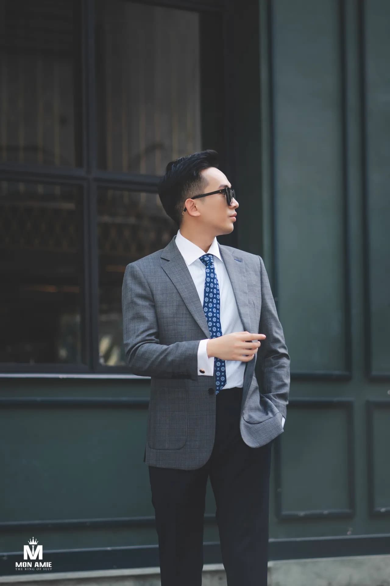 Blazer xám, cà vạt xanh, kính râm, tựa tường cửa hàng xanh rêu