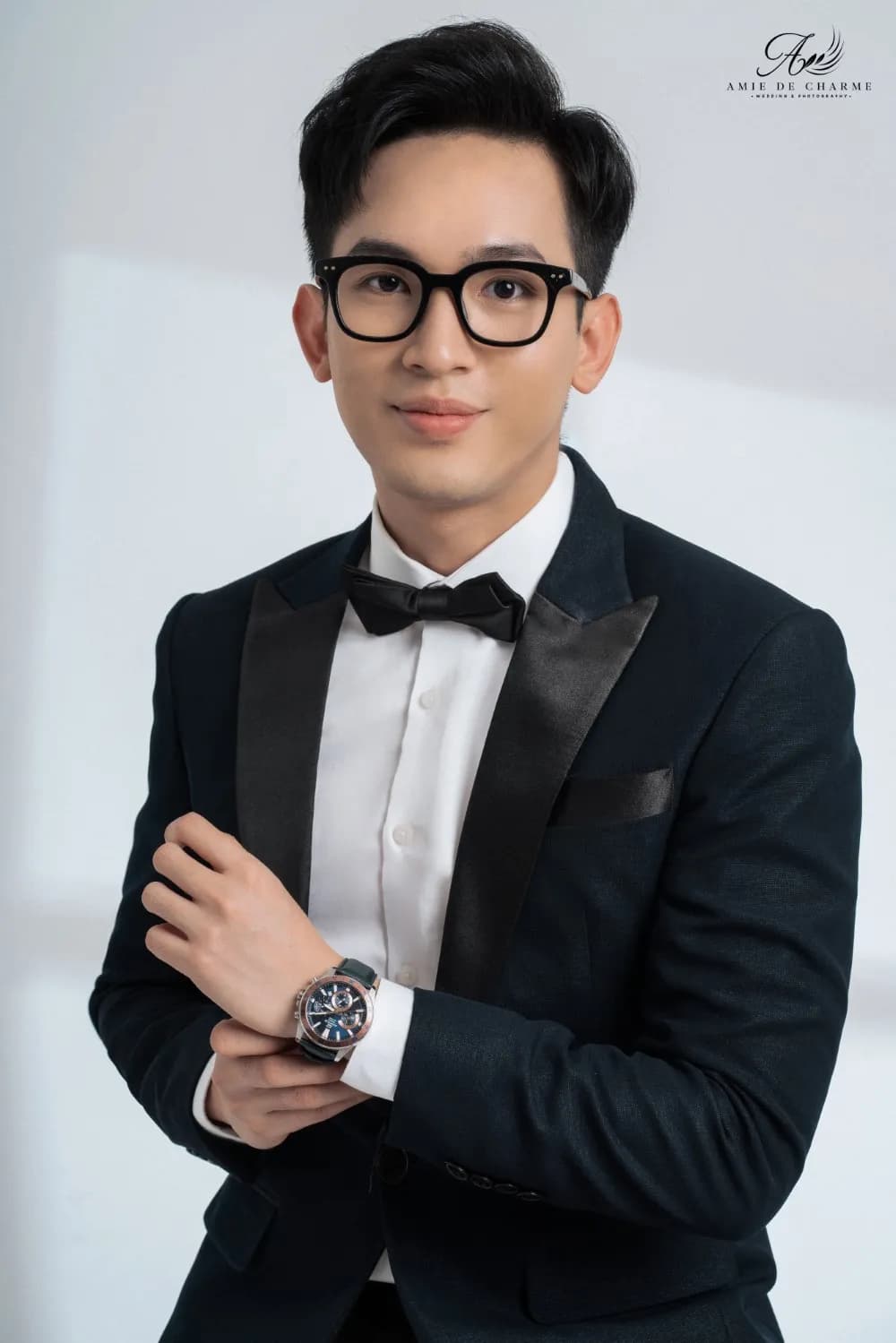 Tuxedo xanh đen cổ ve satin, nơ đen, kính gọng đen, tay chỉnh đồng hồ, chân dung gần