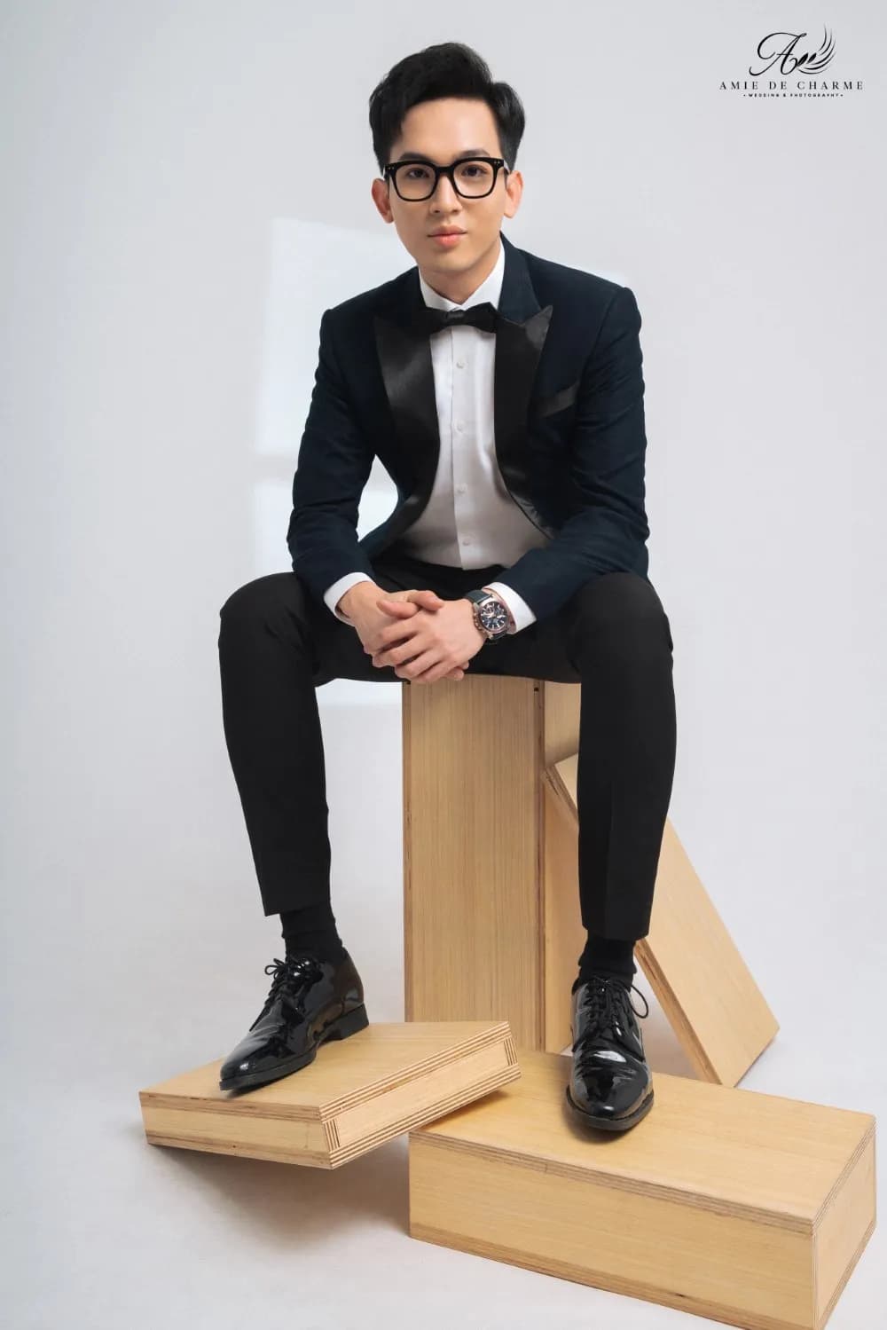 Tuxedo xanh đen, nơ đen, ngồi trên hộp gỗ hai tay đan nhau, giày da bóng