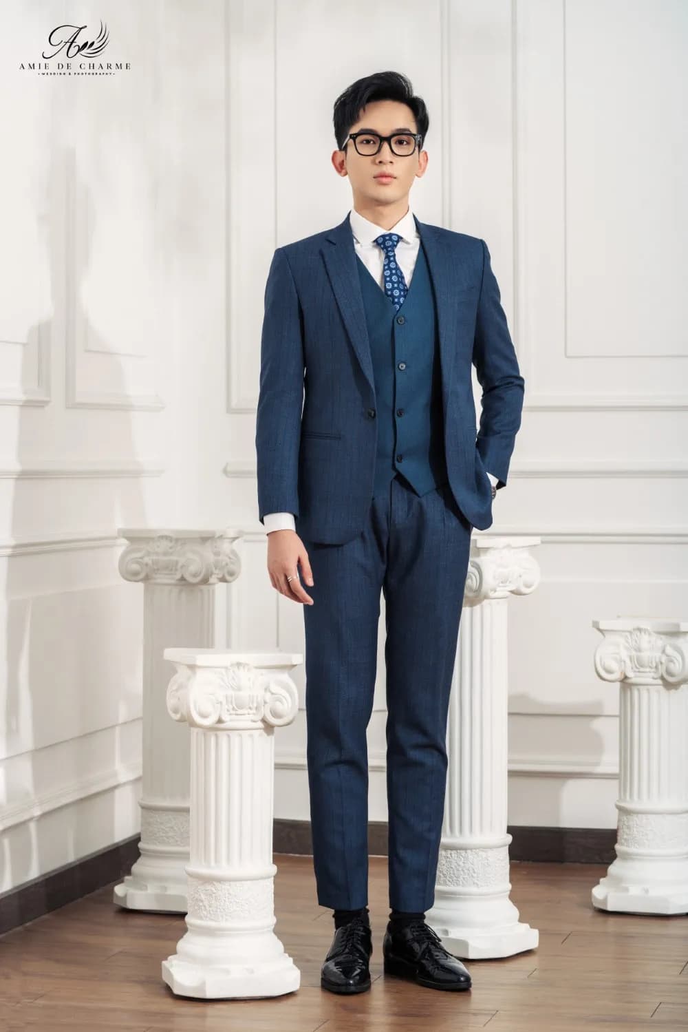 Suit ba mảnh xanh navy kẻ chìm, cà vạt chấm, tay đút túi bên cột La Mã trắng