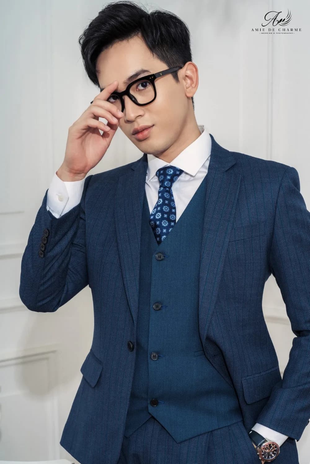 Suit ba mảnh navy, cà vạt chấm hoa, tay chạm gọng kính, chân dung gần