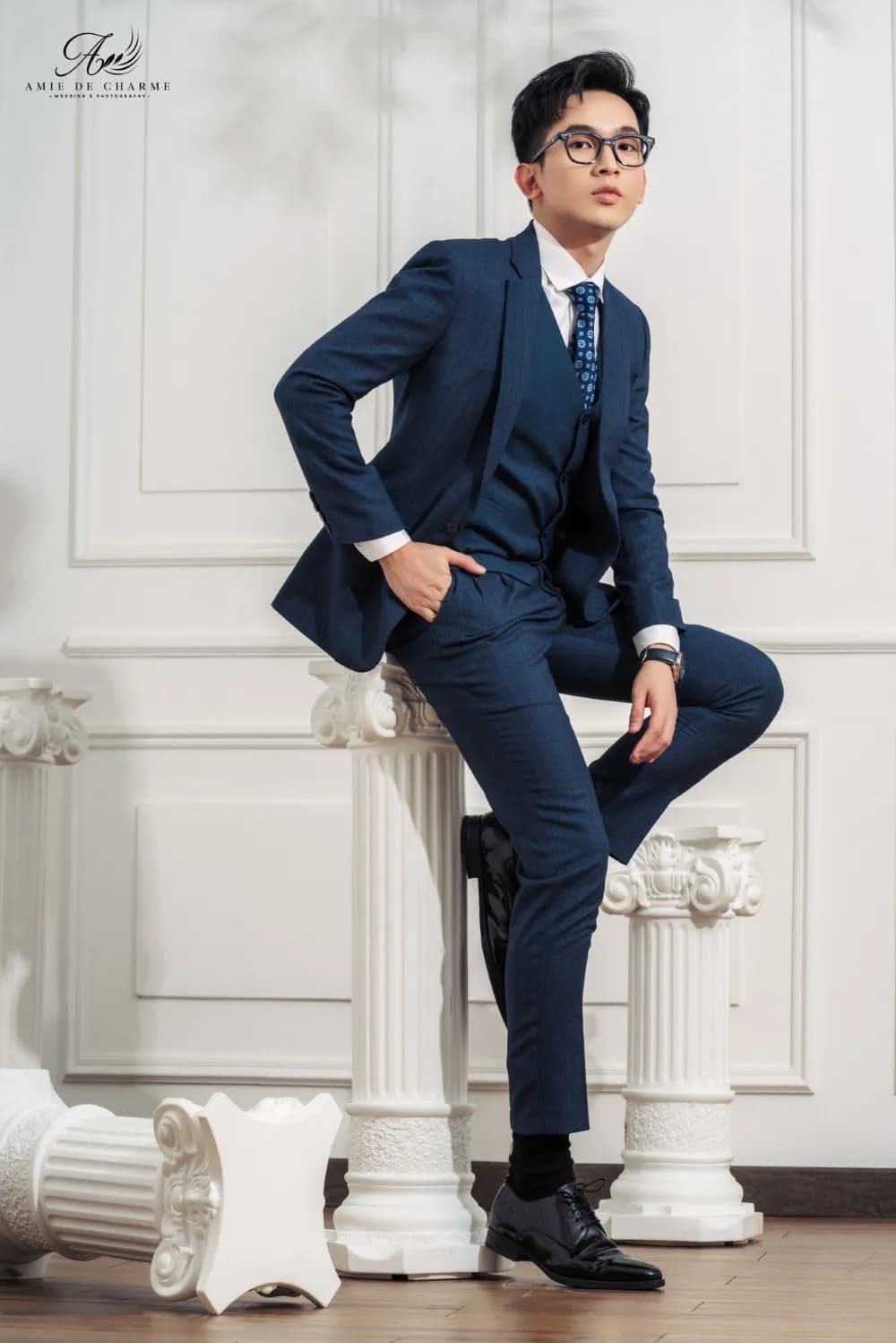 Suit ba mảnh navy, dáng gác chân lên cột La Mã trắng, tay đút túi