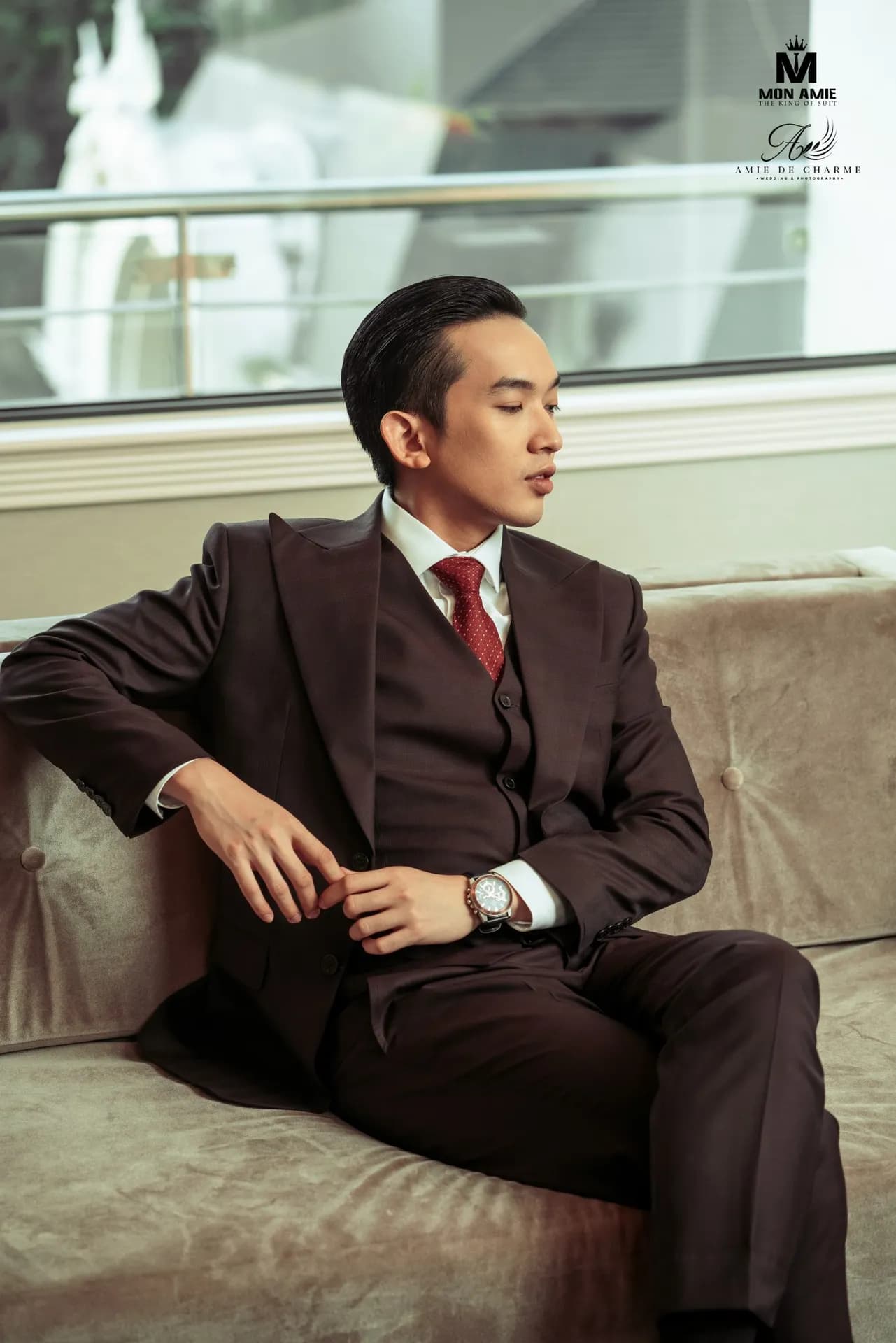 Suit ba mảnh nâu đen, cà vạt đỏ, ngồi tựa sofa nhung, nhìn nghiêng