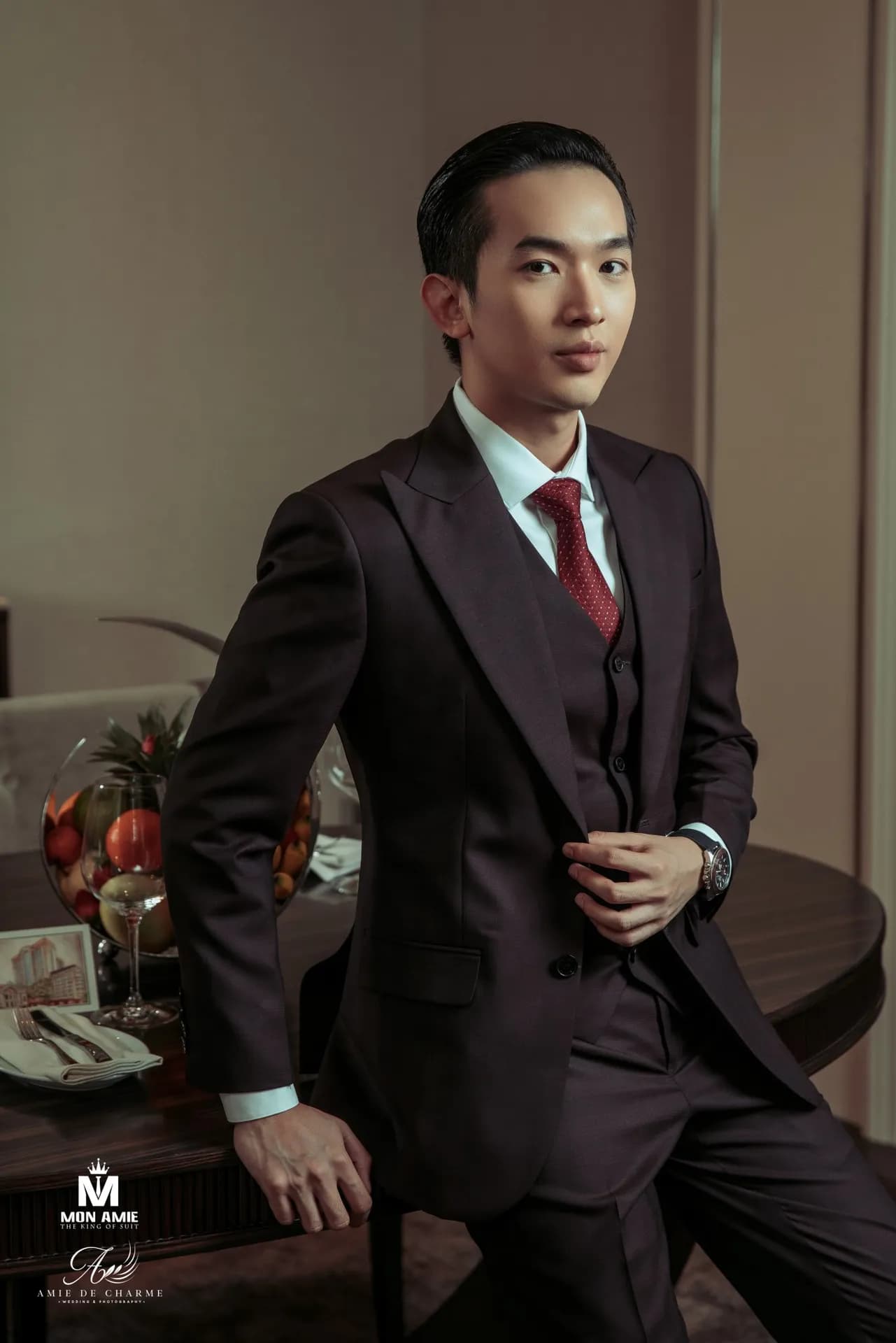 Suit ba mảnh nâu đen, cà vạt đỏ, tựa người chỉnh áo trong phòng ăn ấm