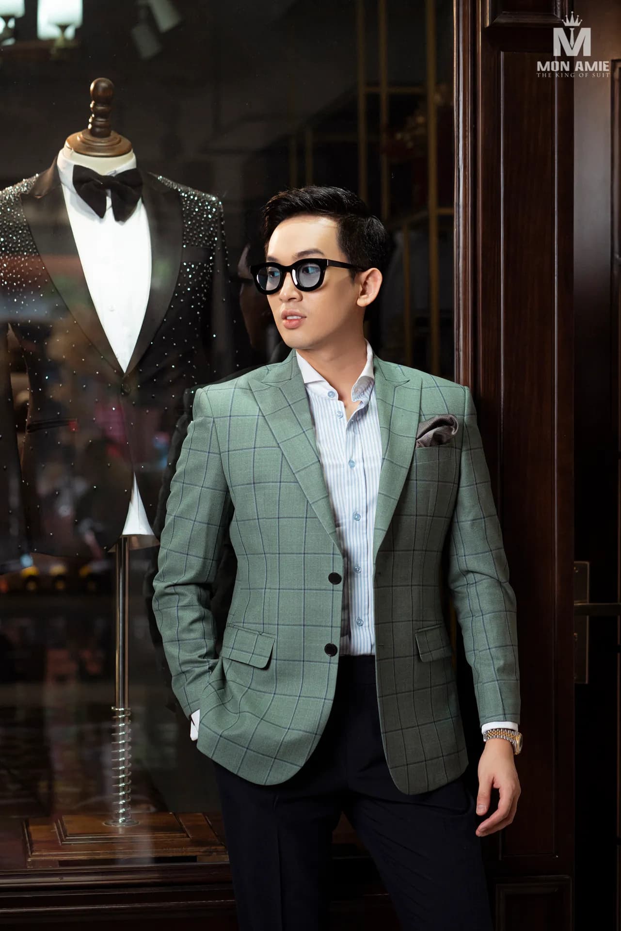 Blazer xanh sage kẻ ô, tay đút túi, cạnh ma-nơ-canh tuxedo kim sa
