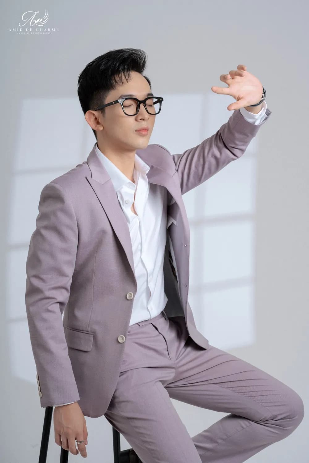 Suit xám tím nhạt, kính đen, ngồi ghế cao tạo dáng trong studio