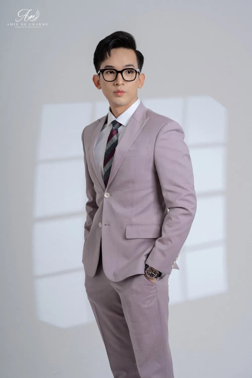 Suit tím hồng nhạt, cà vạt sọc, đứng tay đút túi, nhìn chéo, nền studio xám