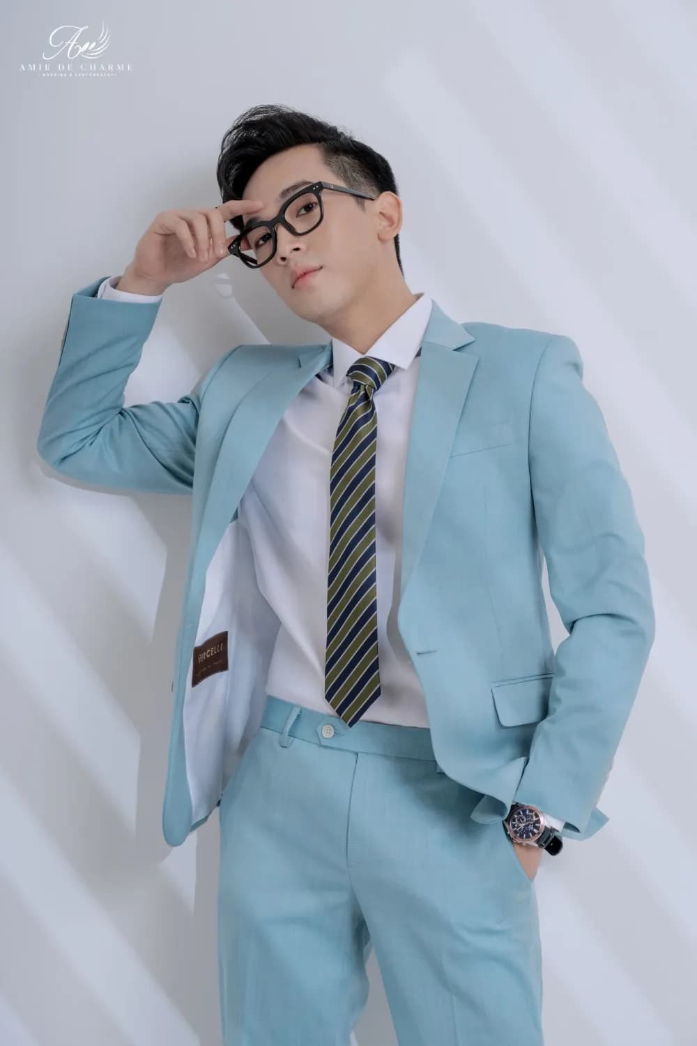 Suit xanh ngọc nhạt, tựa tường, tay đẩy kính, lộ lớp lót áo