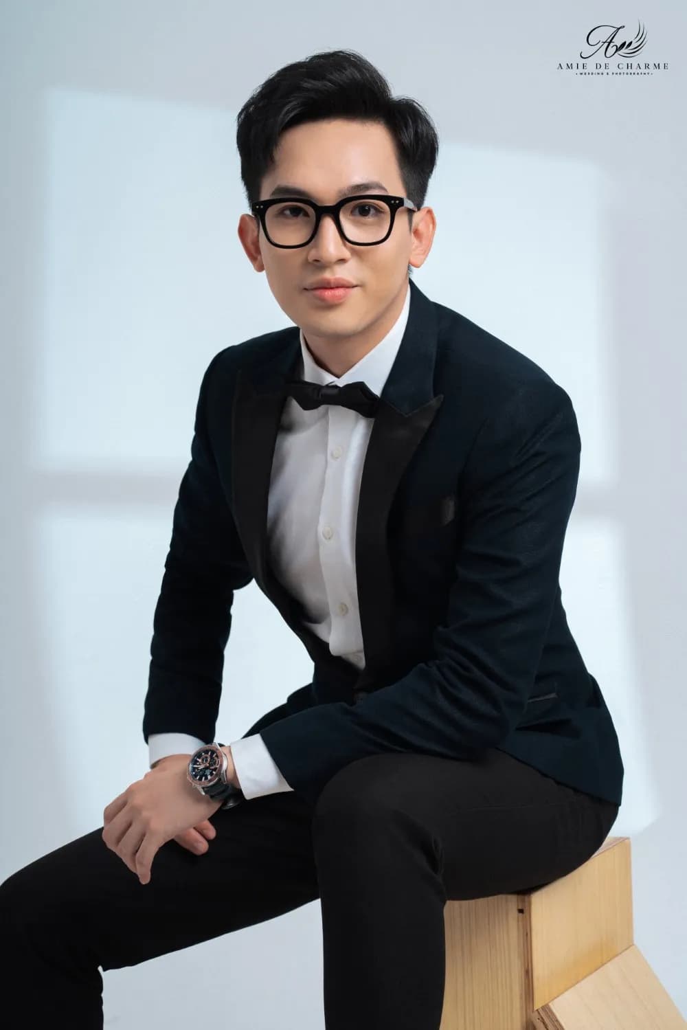 Tuxedo xanh đen, nơ đen, ngồi nghiêng người trên hộp gỗ nền studio sáng