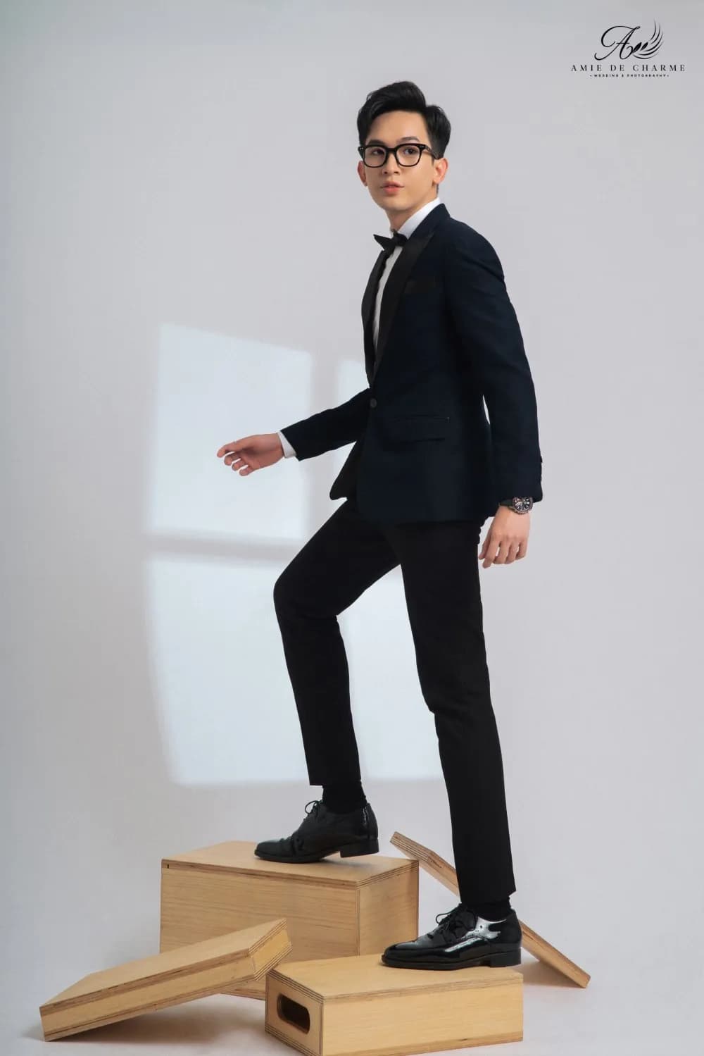 Tuxedo xanh đen, nơ đen, bước chân lên hộp gỗ, dáng năng động nền studio