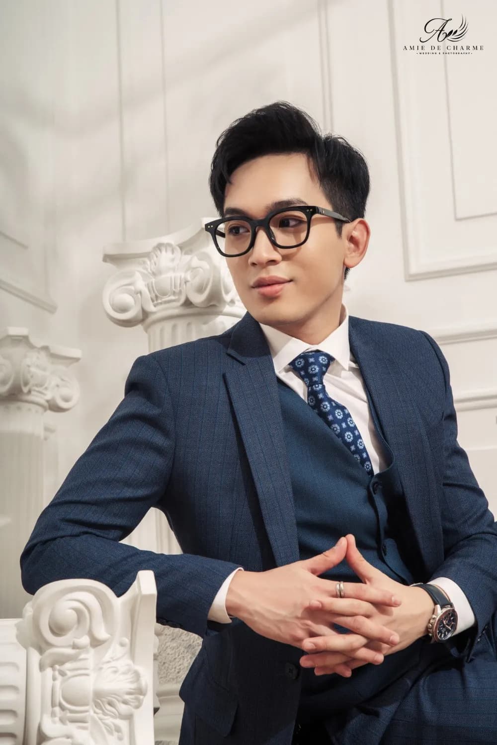 Suit ba mảnh navy, hai tay đan nhau, ngồi cạnh cột La Mã trắng