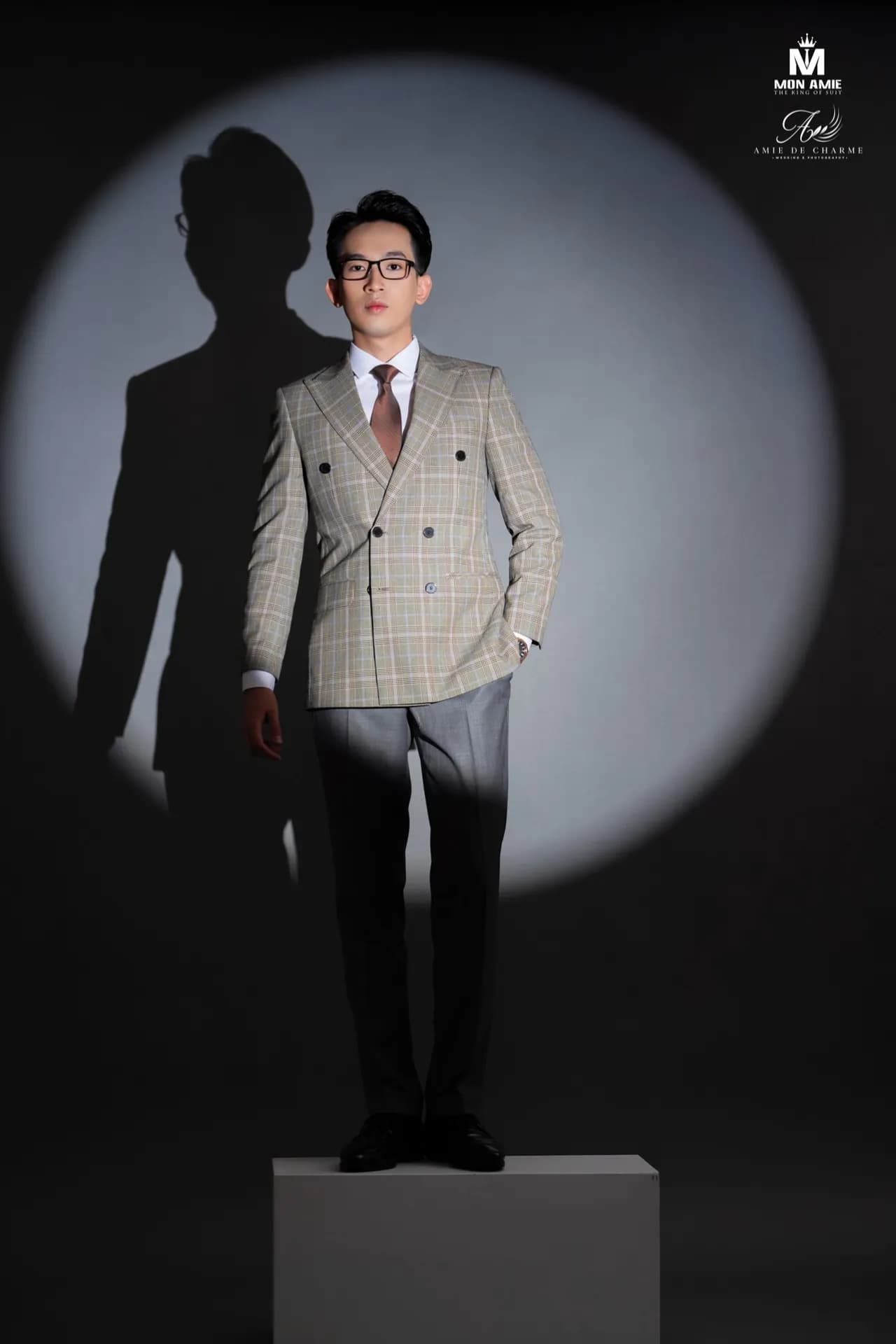 Suit cài hai hàng be kẻ ô, quần xám, đứng trên bục giữa vòng sáng spotlight