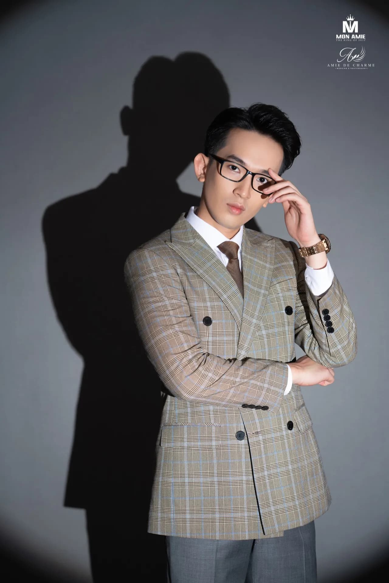 Suit cài hai hàng be kẻ ô, tay chạm gọng kính trong vòng sáng spotlight