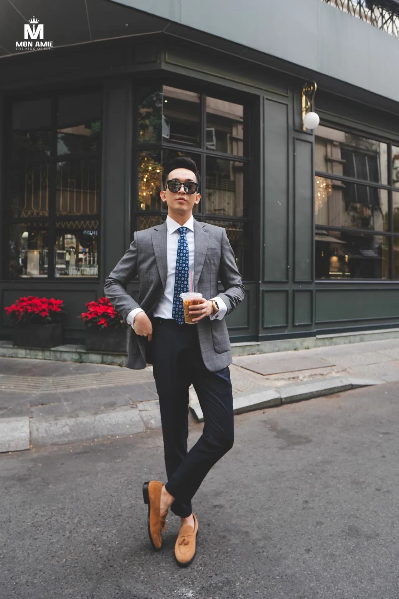 Blazer xám, cà vạt xanh, cầm ly cà phê, sải bước trước cửa hàng xanh rêu