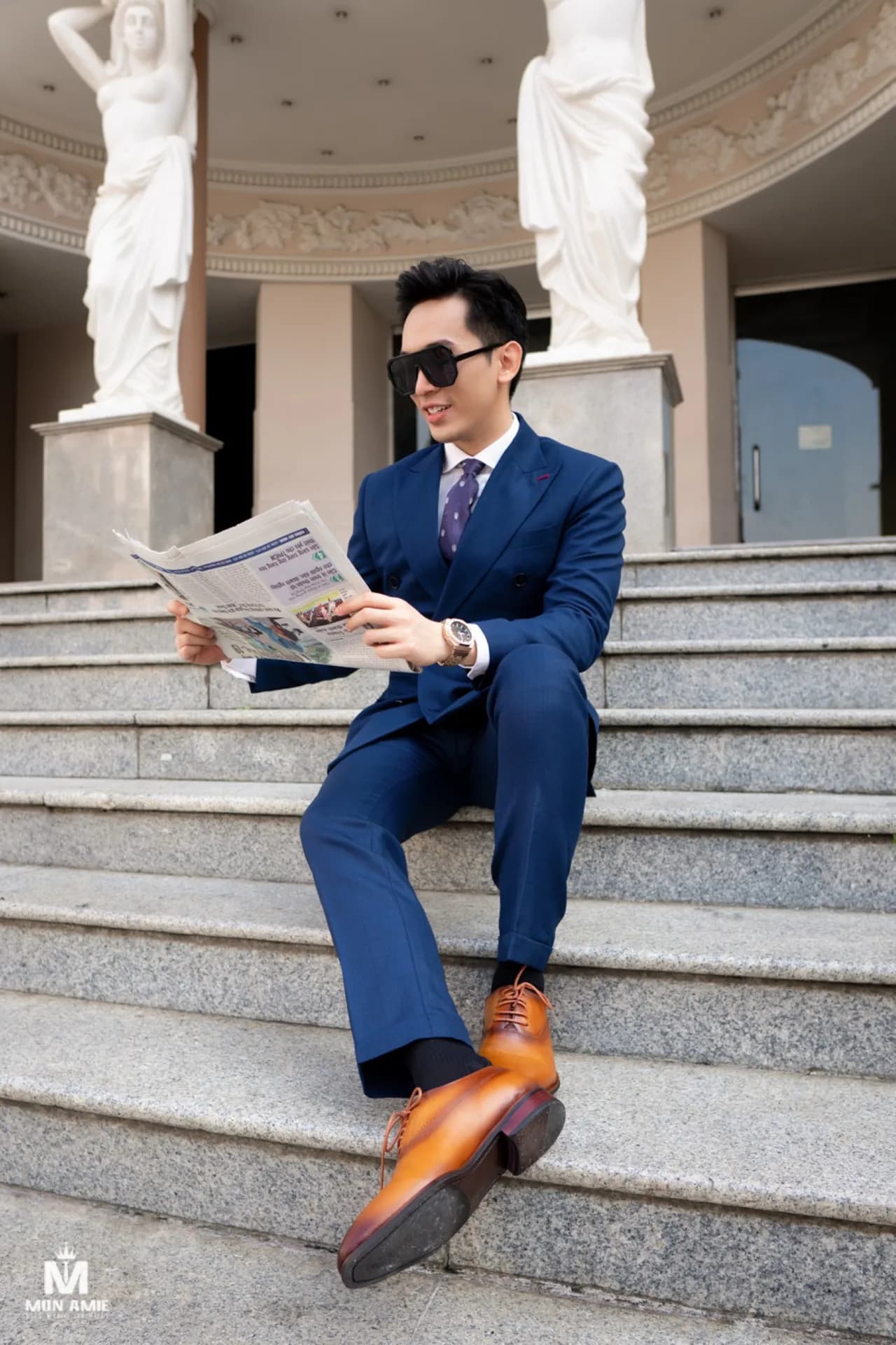 Suit xanh royal ngồi bậc thềm đá, giày da nâu, tay cầm tờ báo