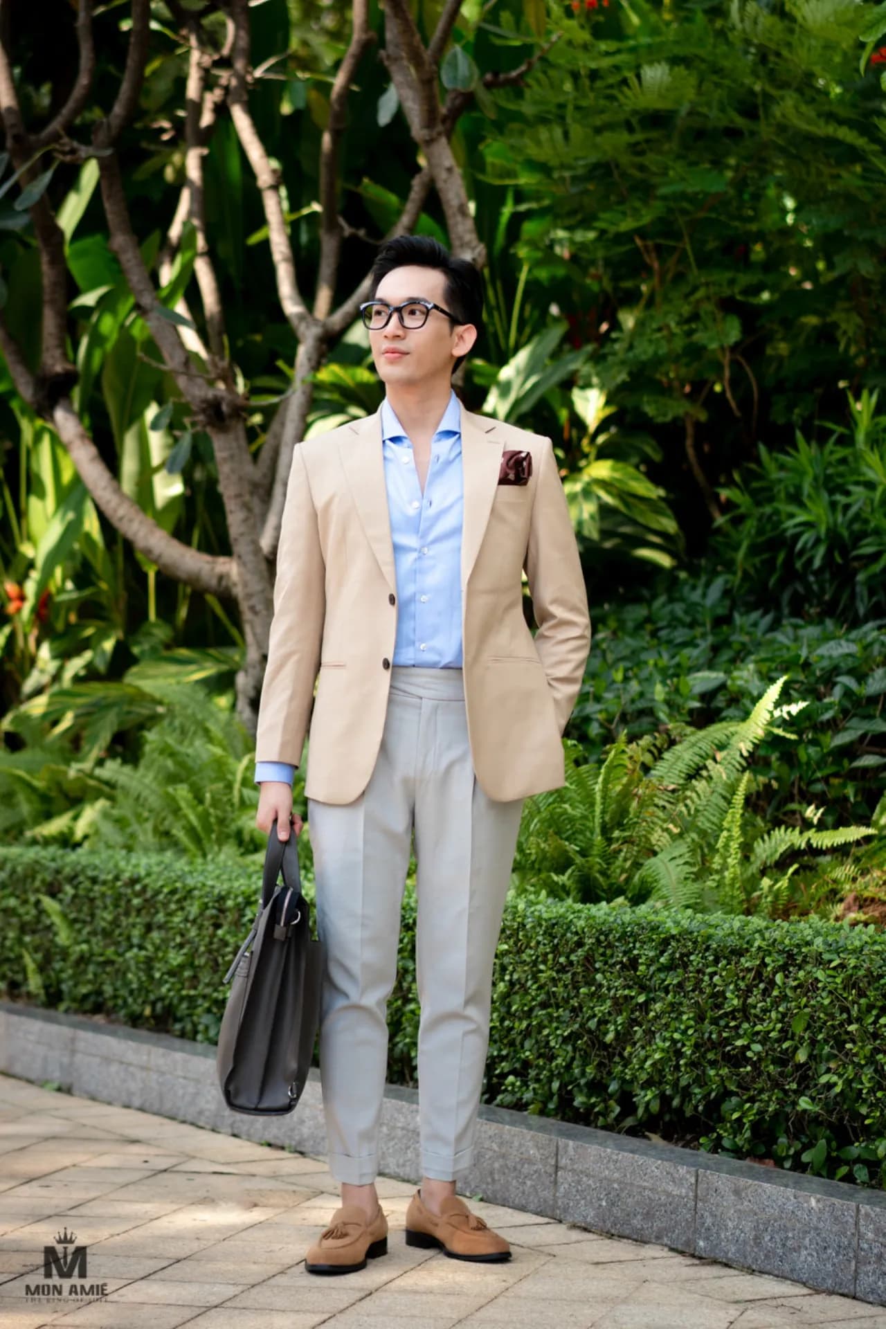 Blazer be, sơ mi xanh nhạt, quần sáng, cầm cặp da, nền cây xanh