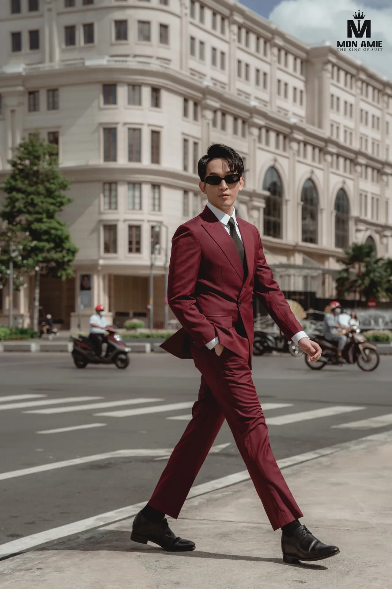Suit đỏ đô, kính râm, sải bước qua phố, nền tòa nhà kem