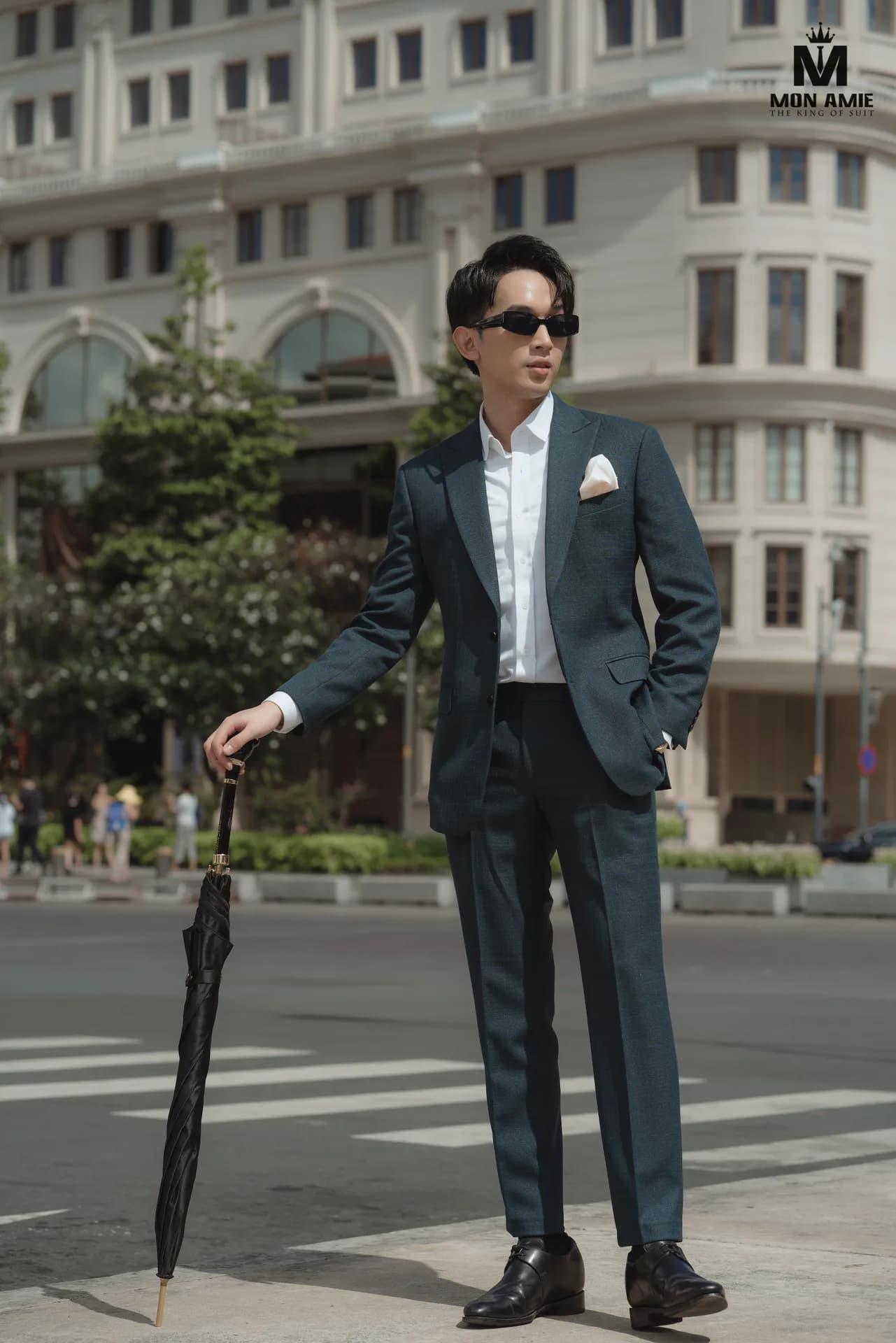 Suit xanh rêu, kính râm, tay cầm ô gập, đứng giữa phố