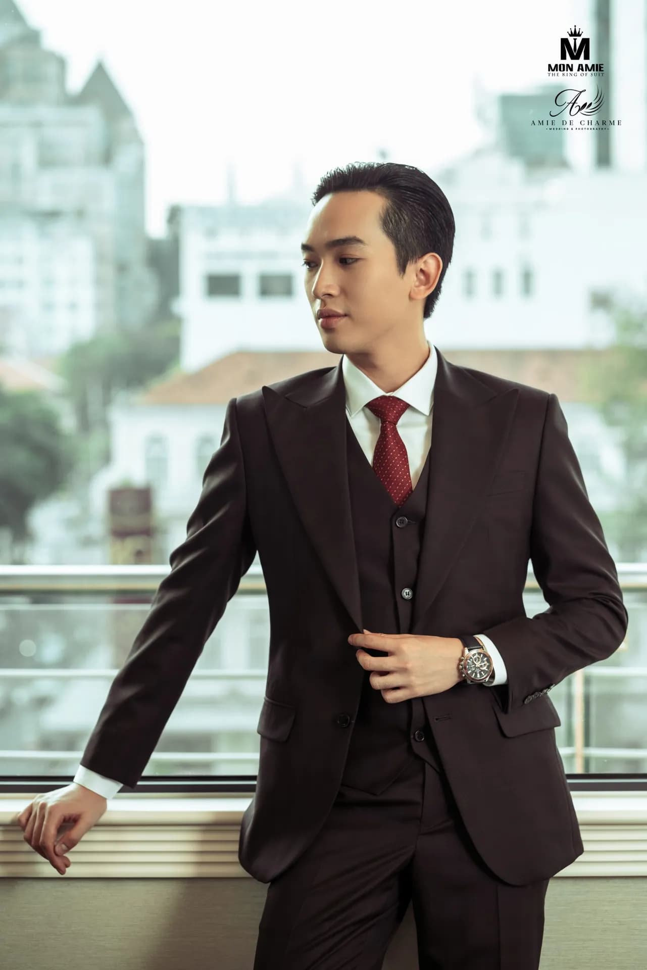 Suit ba mảnh nâu đen, cà vạt đỏ, tựa lan can cạnh cửa sổ nhìn xa