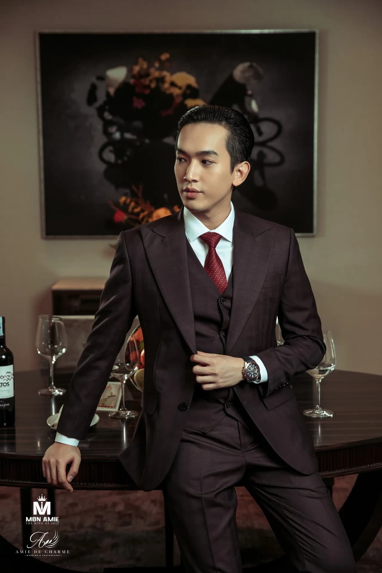 Suit ba mảnh nâu đen, cà vạt đỏ, tựa bàn ăn nhìn nghiêng trước bức tranh
