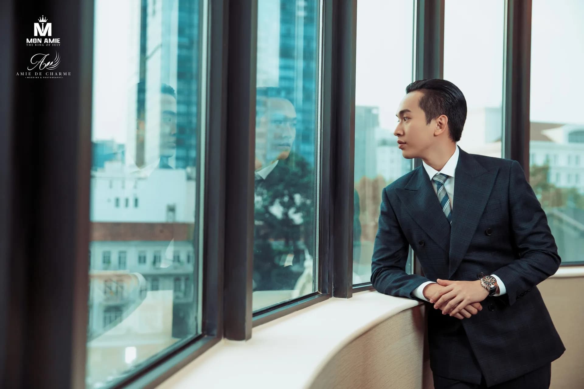 Suit navy cài hai hàng, khoanh tay tựa bệ cửa sổ nhìn ra thành phố