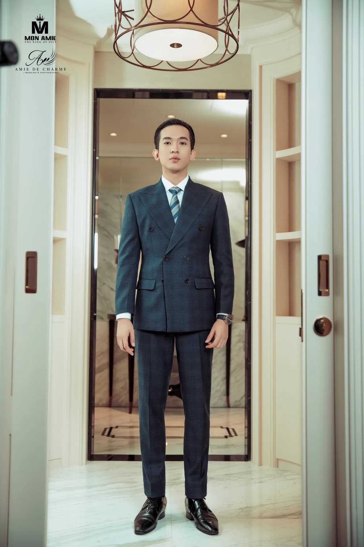 Suit navy cài hai hàng, cà vạt sọc, đứng thẳng giữa hành lang sảnh khách sạn