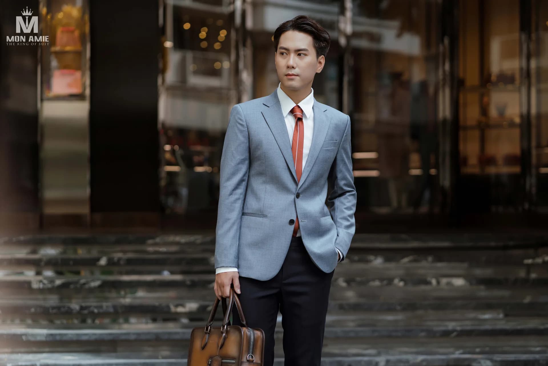 Suit xám xanh, cà vạt cam và cặp da trước mặt tiền đá