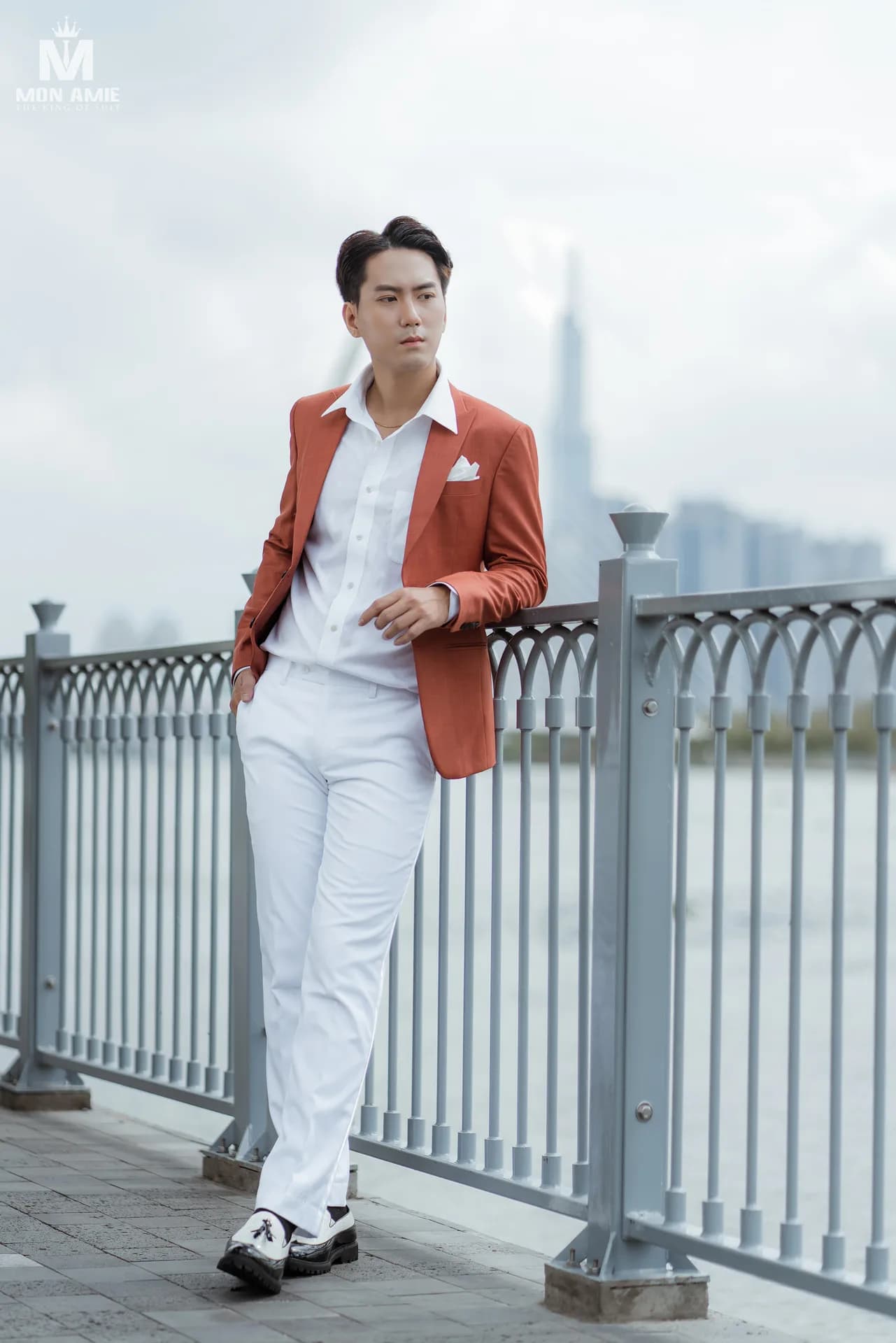 Blazer cam đất và quần trắng tựa lan can ven sông