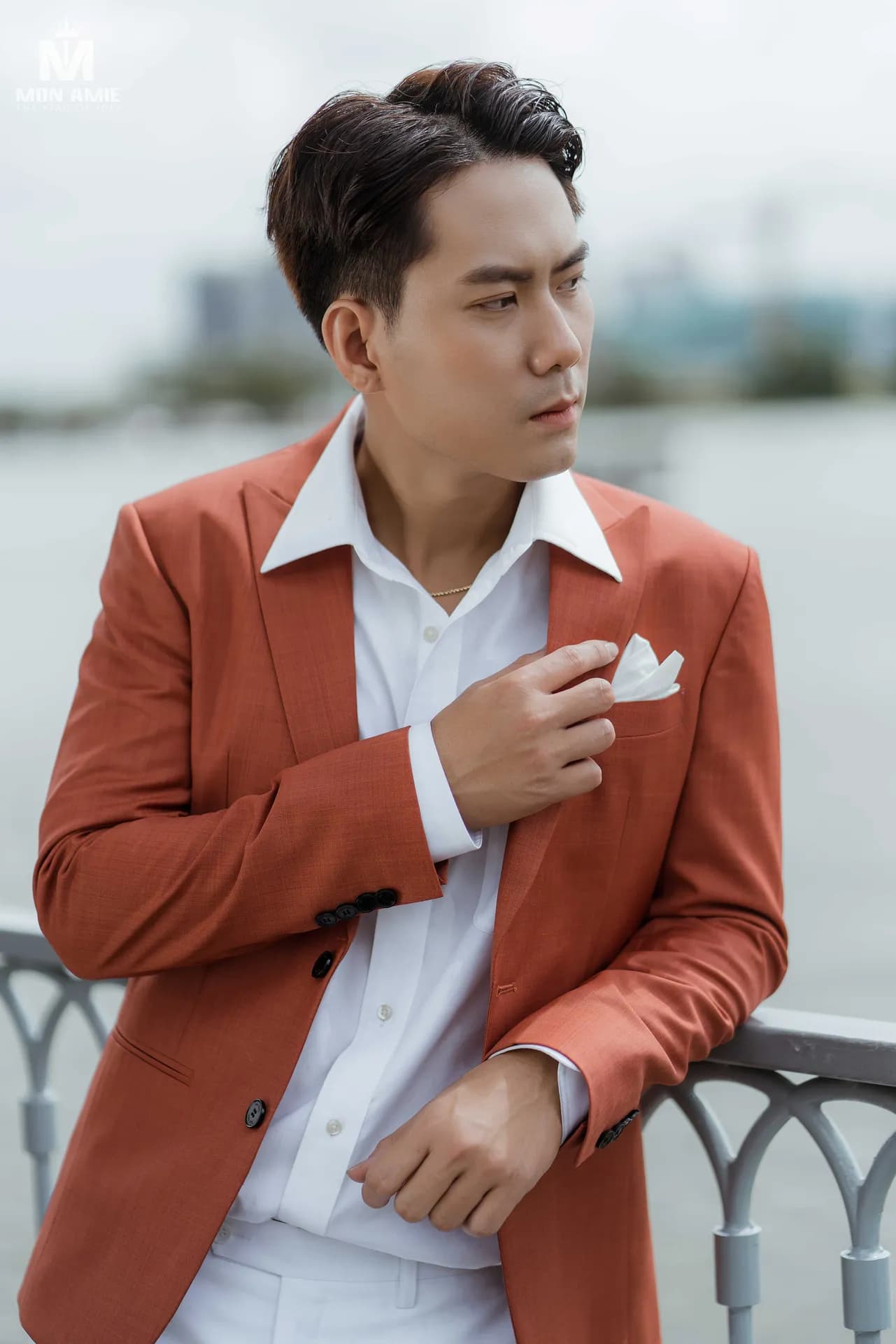 Cận cảnh vest cam đất, khăn cài túi trắng, dựa lan can bên sông