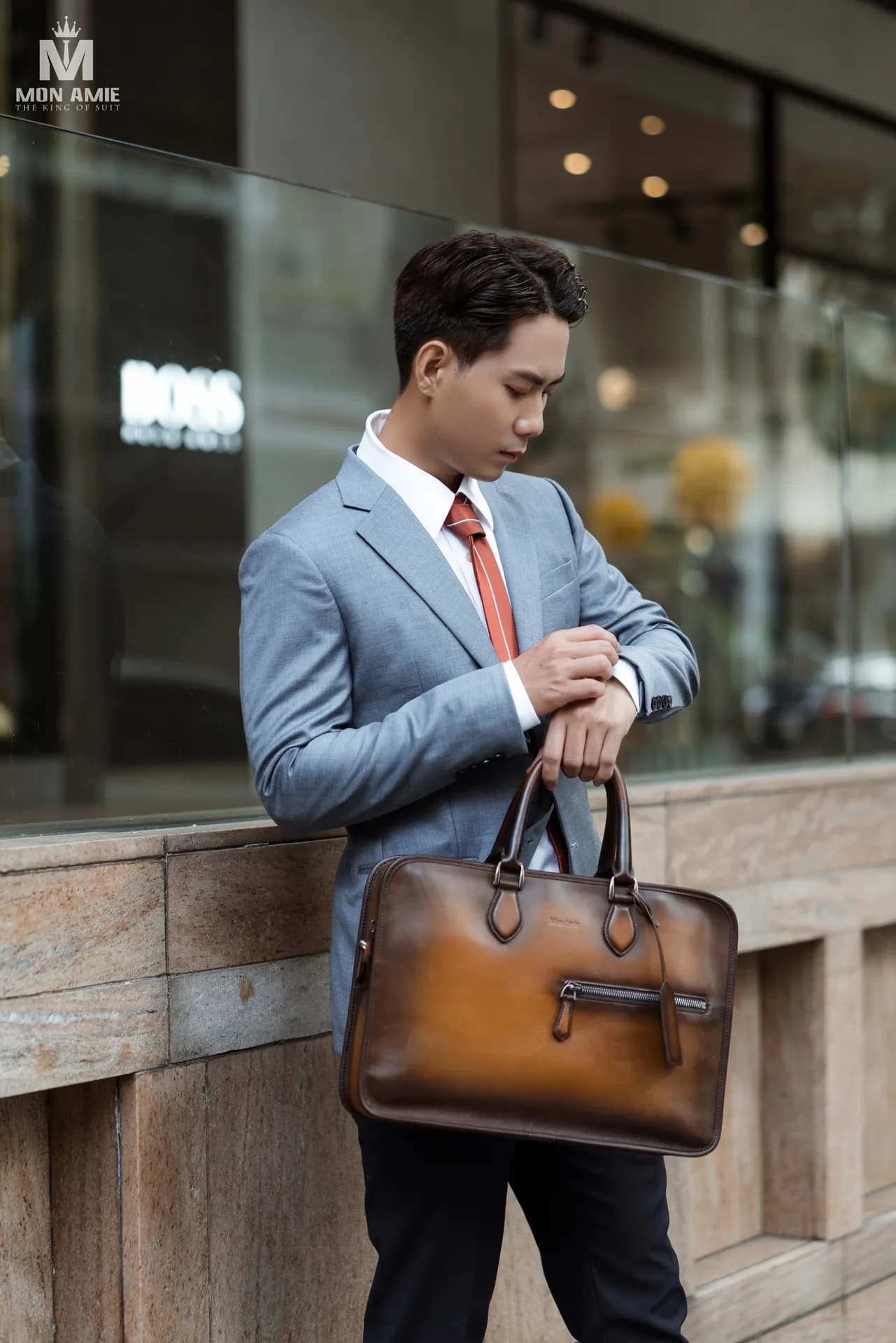 Vest xám xanh, cà vạt cam, xách túi da nâu, dựa tường đá