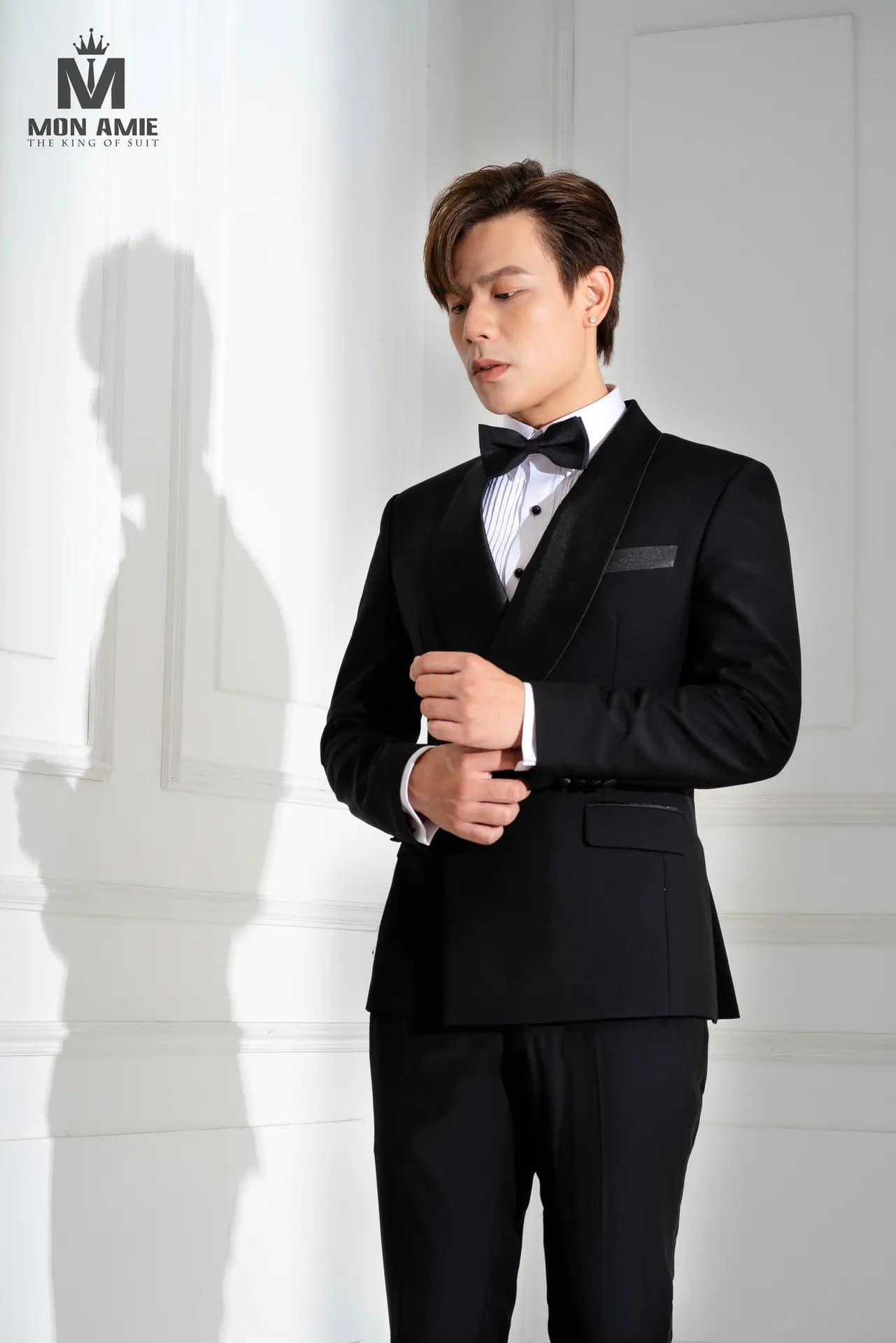 Tuxedo đen nơ, nghiêng mặt, chỉnh măng sét bên bức tường trắng