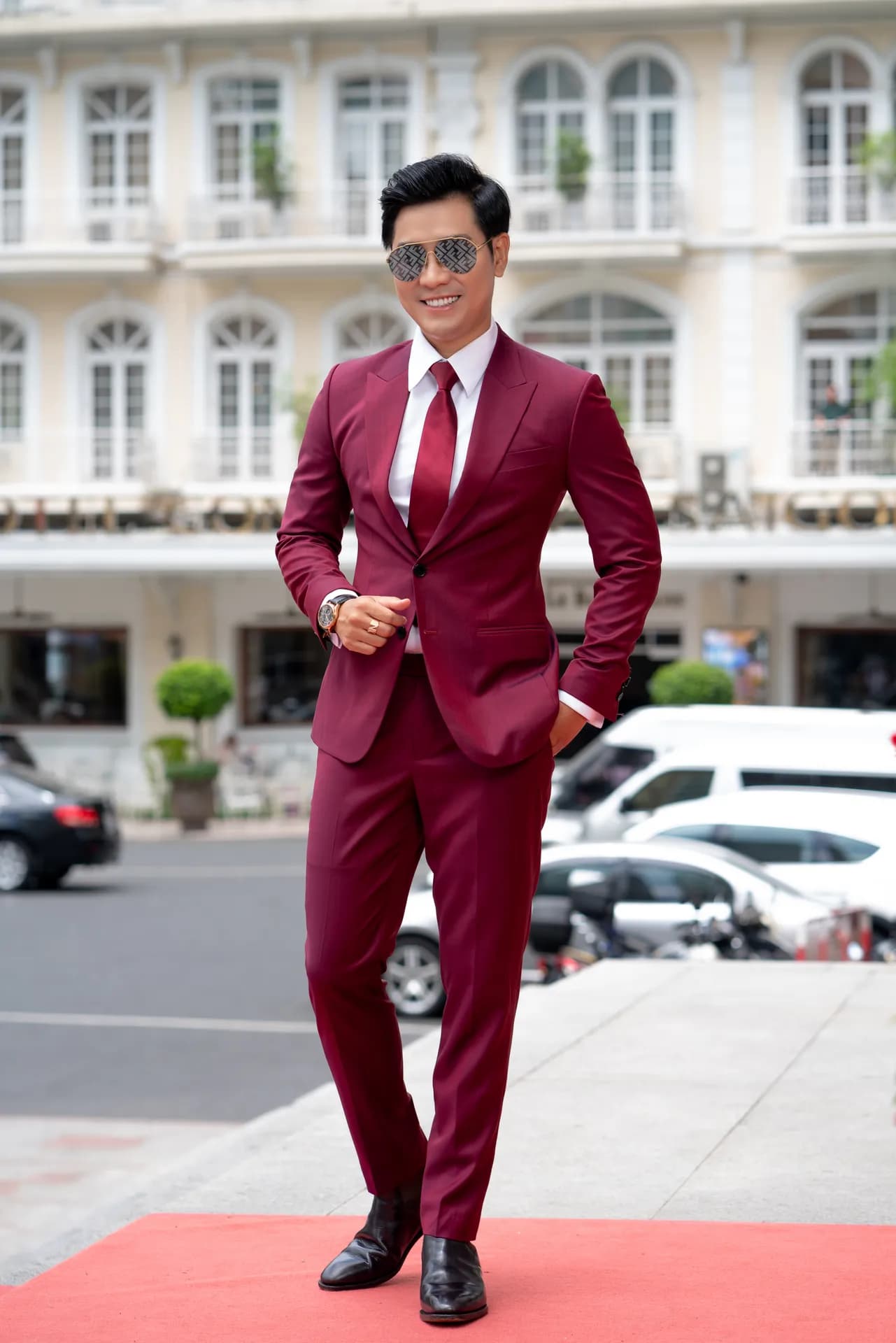 Suit đỏ đô, kính râm, bước trên thảm đỏ trước tòa nhà phố cổ