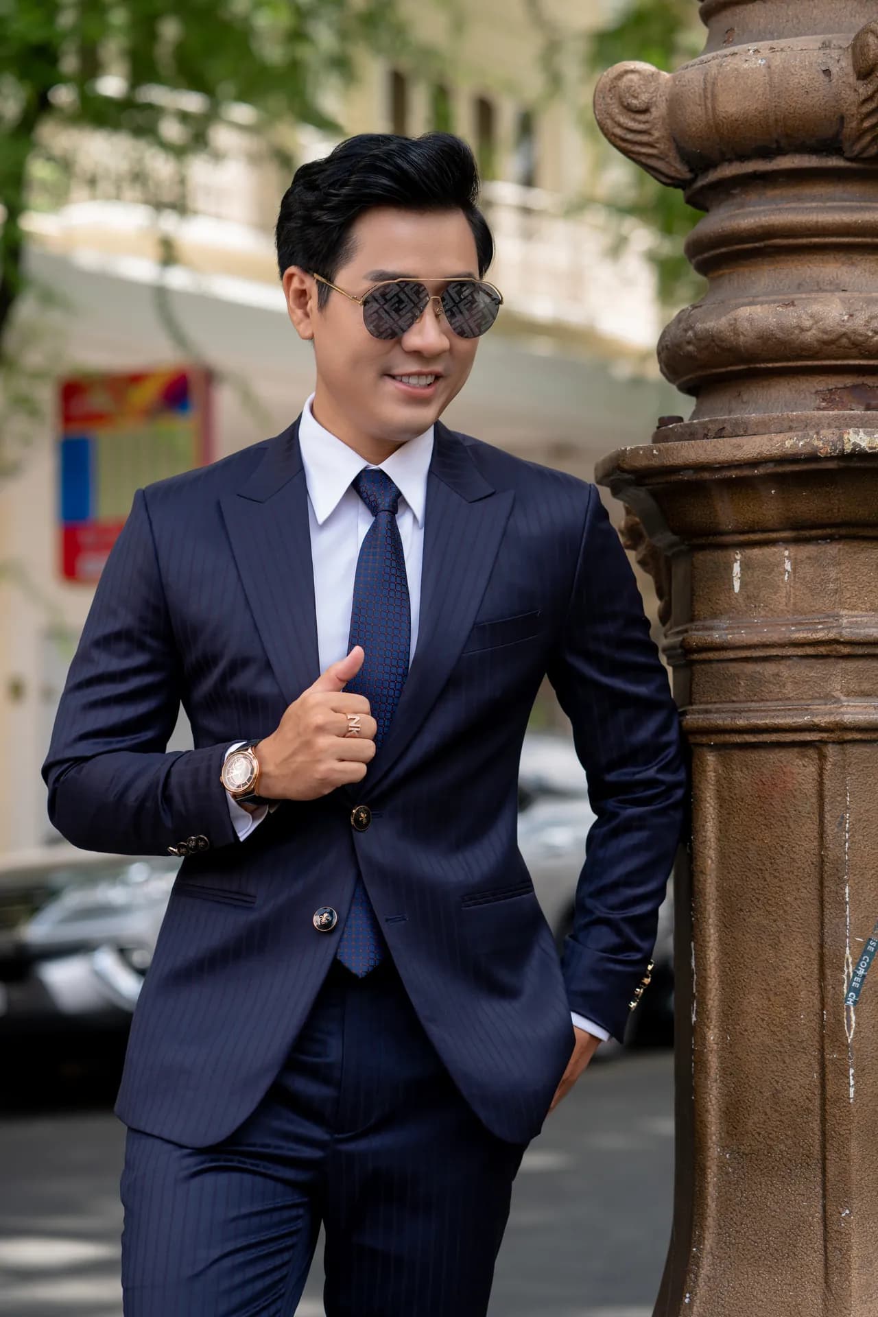 Suit xanh navy kẻ sọc, kính râm, tay cài khuy áo, dựa cột đá