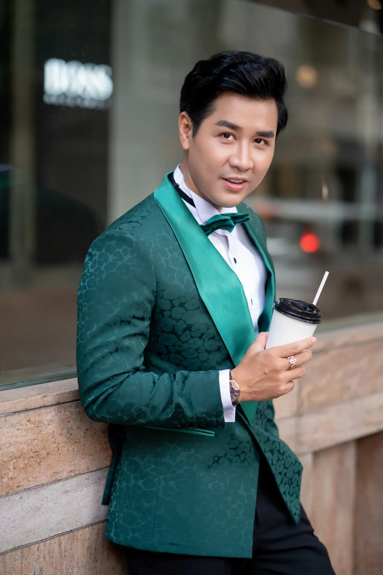 Tuxedo nhung xanh lục cổ shawl, nơ đen, tựa tường đá, cầm ly cà phê