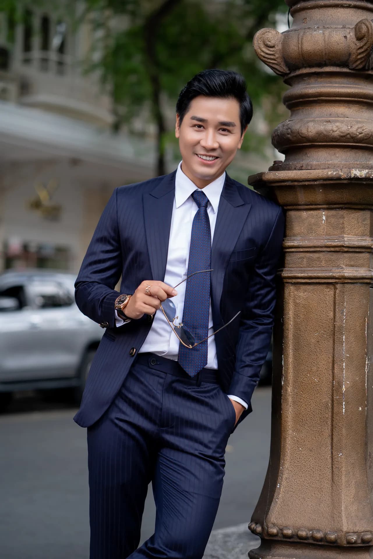 Suit xanh navy kẻ sọc, cà vạt xanh, dựa cột đá trên phố