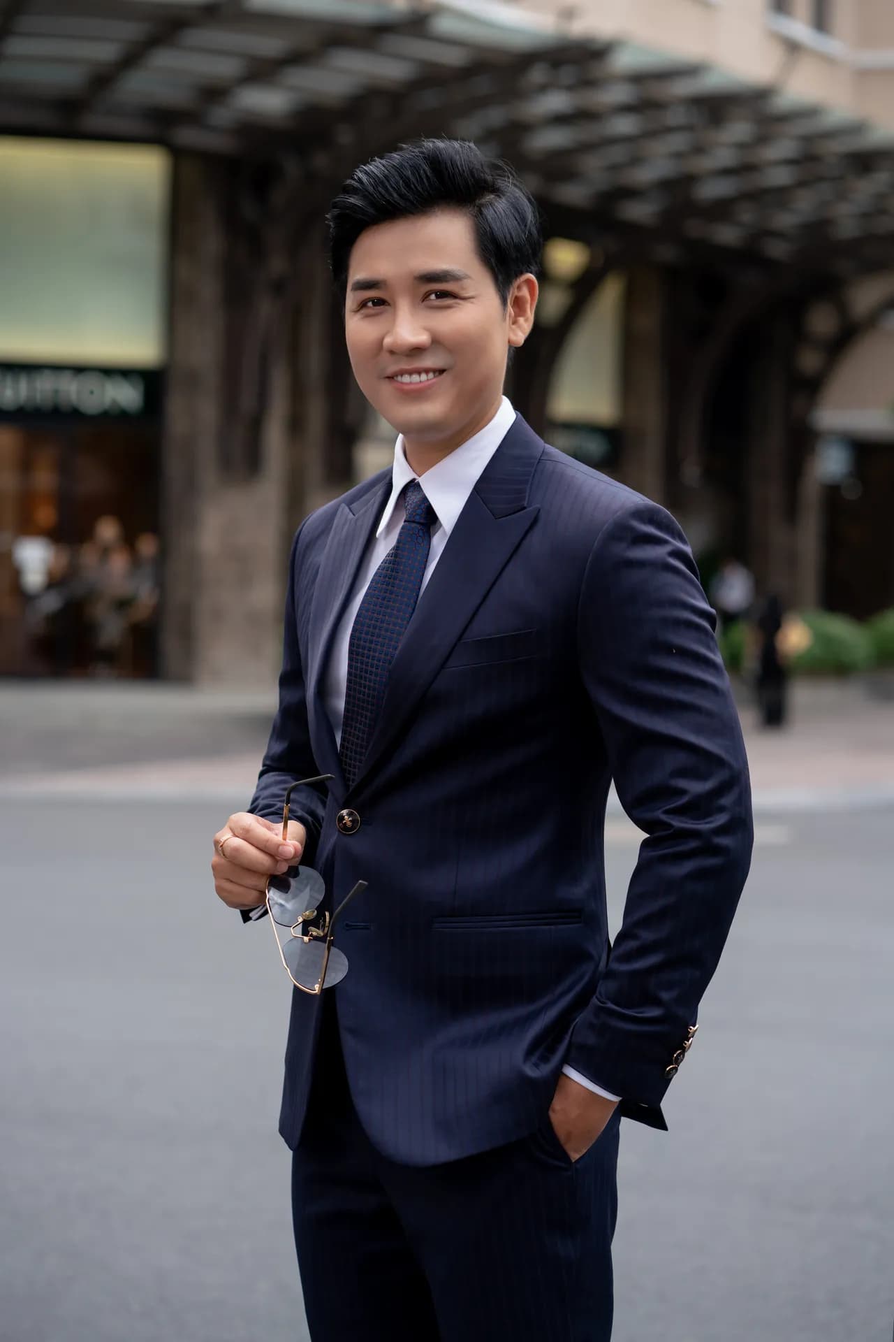 Blazer xanh navy, sơ mi trắng, tay đút túi, mỉm cười trên phố