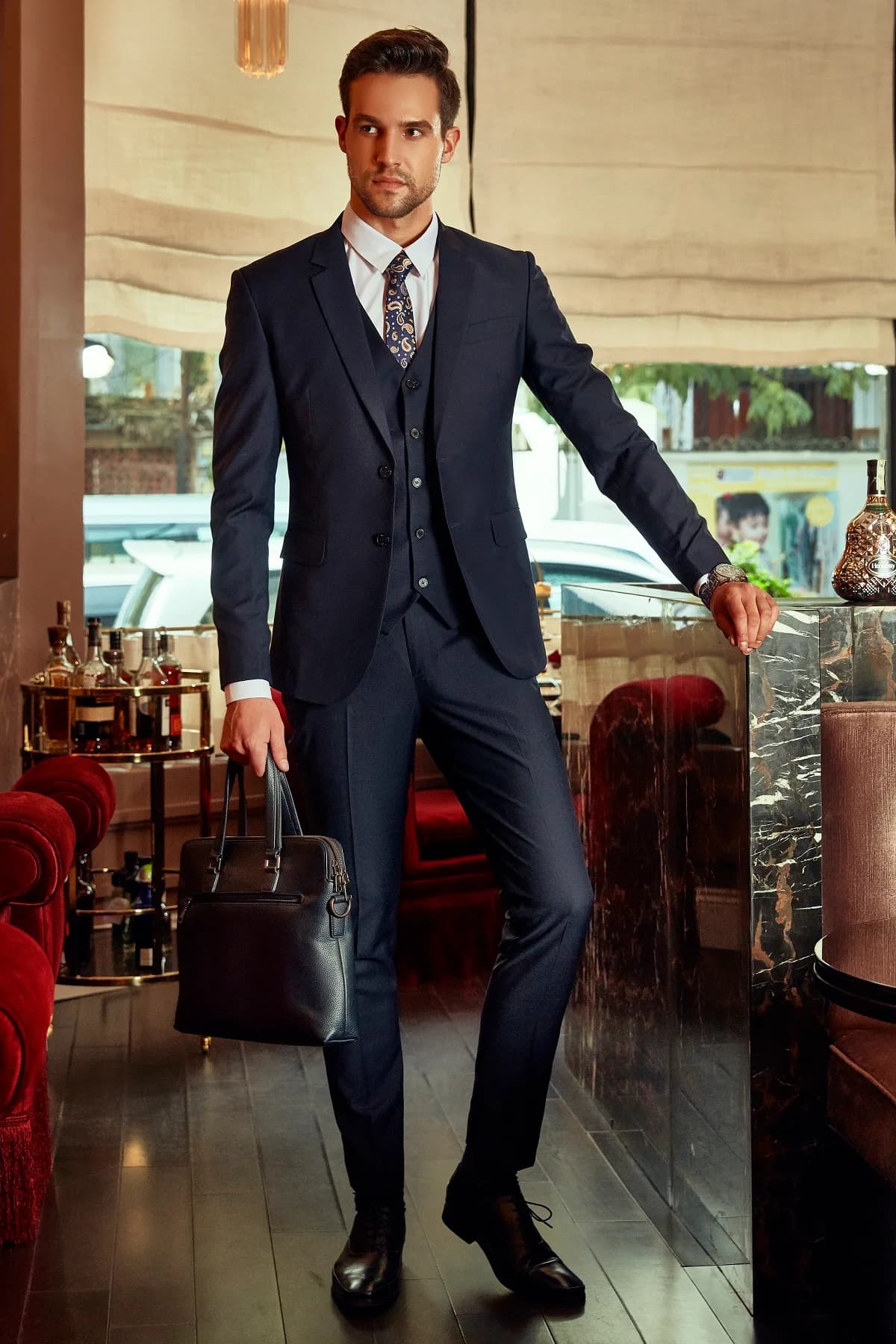 Suit ba mảnh xanh navy, cà vạt paisley, cặp da đen, tựa quầy bar đá cẩm thạch