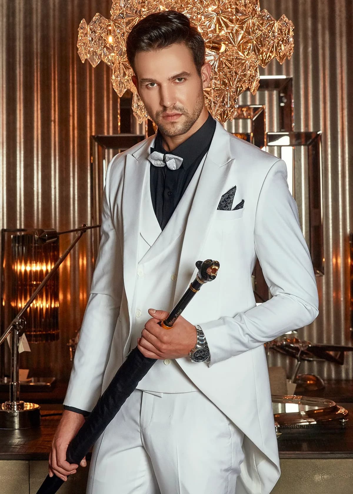 Tailcoat trắng ba mảnh, nơ bạc, cầm gậy dưới đèn chùm pha lê
