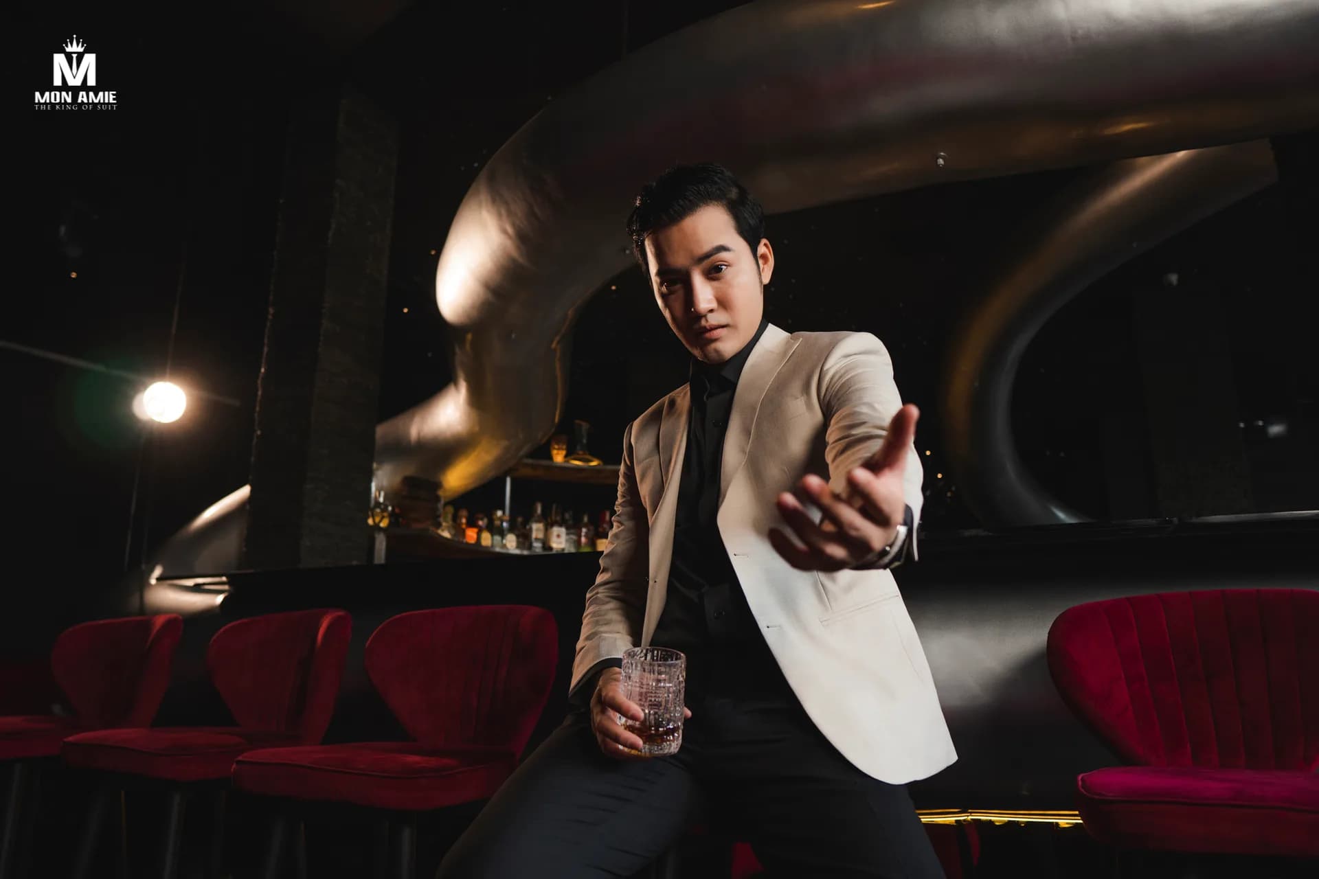 Suit ghi ba mảnh, nơ cổ và ghim cài, dáng ngồi ghế gỗ trong studio