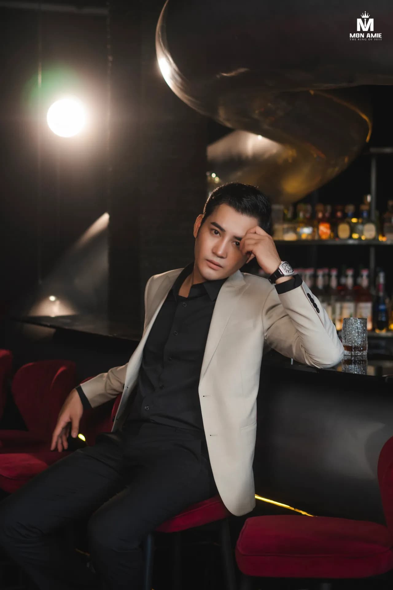 Vest kem, sơ mi đen, tựa tay bên quầy bar ánh đèn ấm