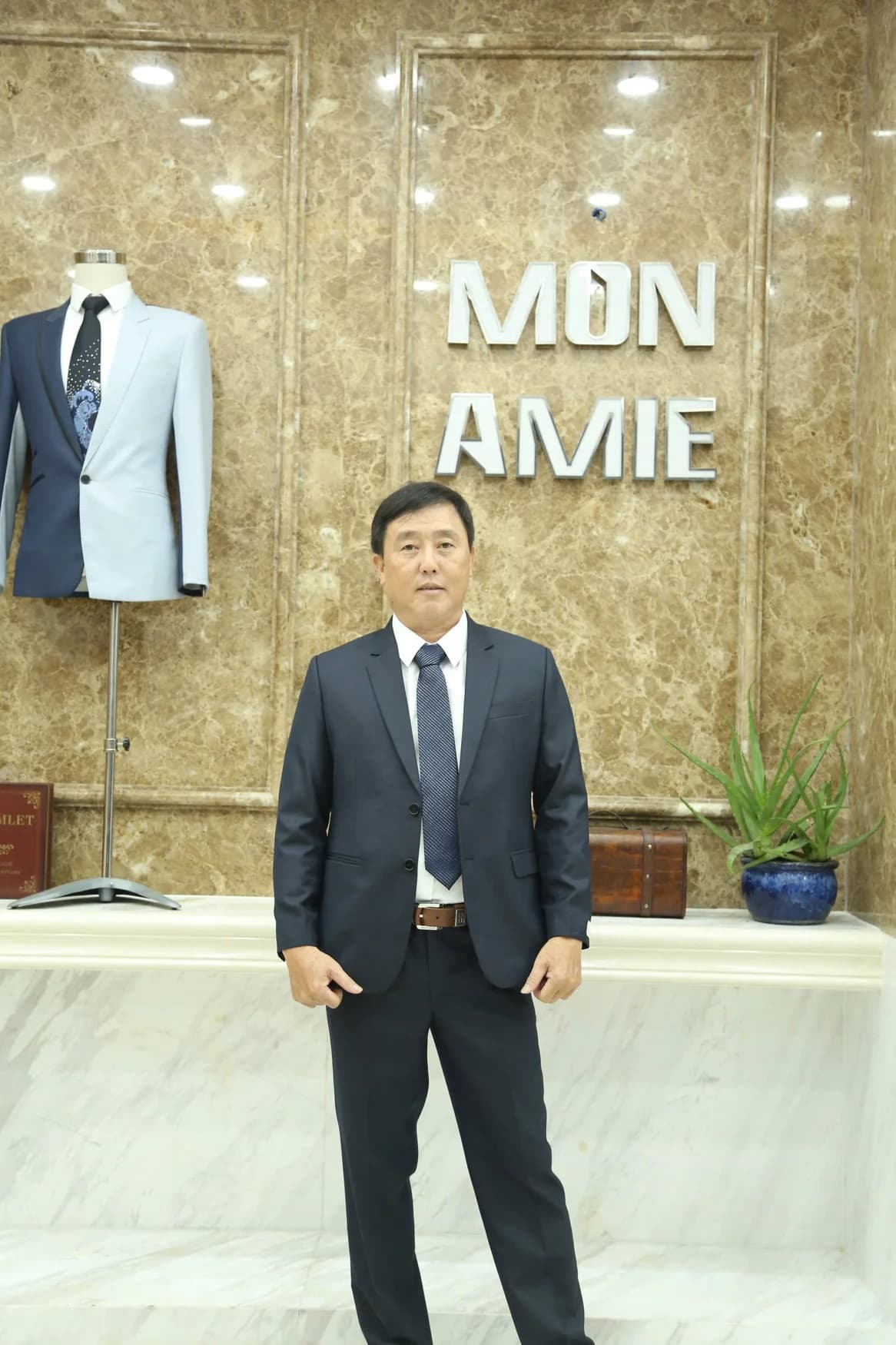 Quý ông vest navy đậm, cà vạt xanh, nền logo Mon Amie