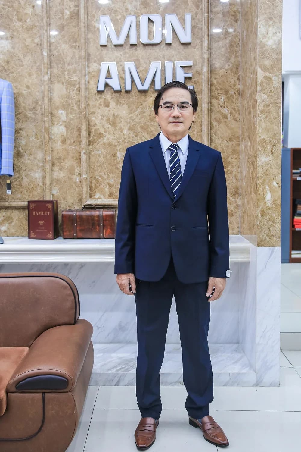 Suit xanh navy cà vạt kẻ, kính trí thức, đứng trước biển hiệu Mon Amie