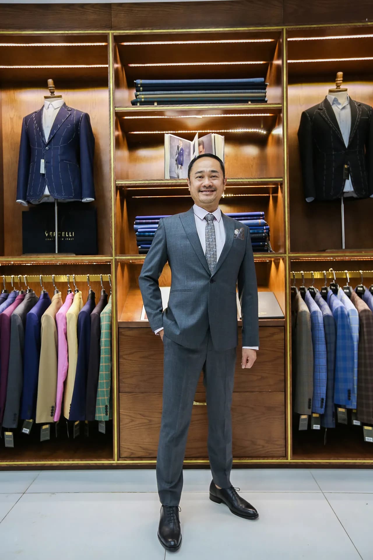 Suit xám ánh xanh, cà vạt họa tiết paisley, nụ cười rạng rỡ giữa tủ vải gỗ ấm