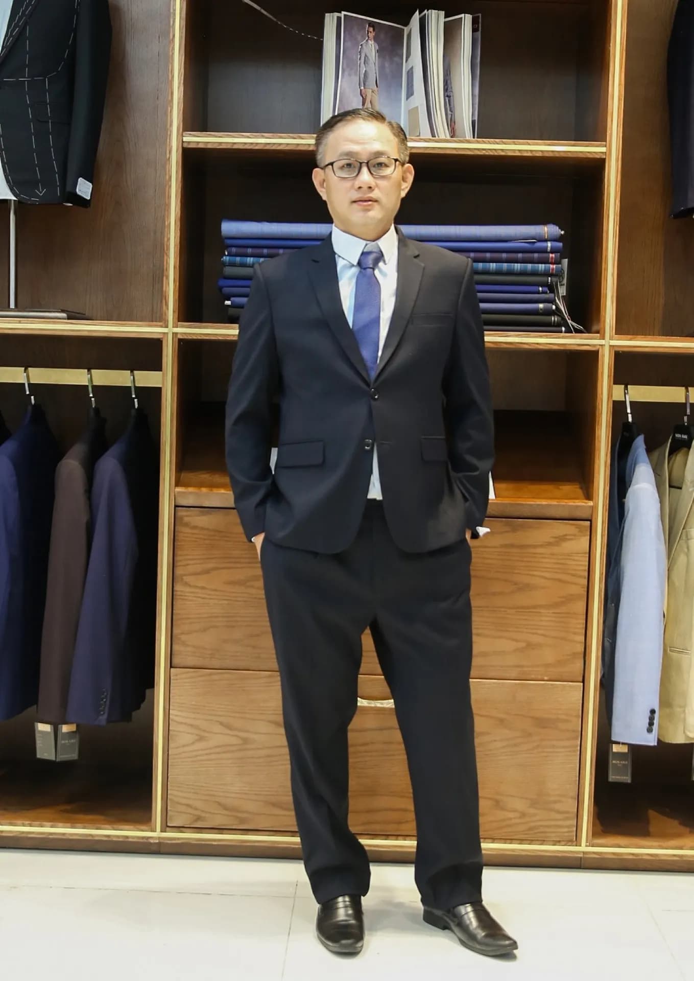 Suit navy hai cúc, cà vạt xanh, sơ mi xanh nhạt trước kệ vải gỗ tinh tế