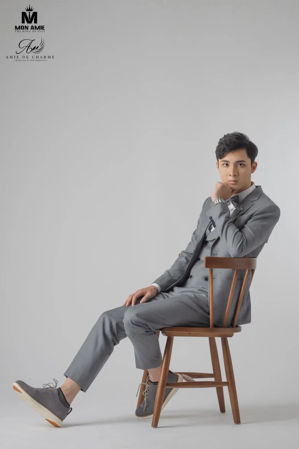 Suit xám ba mảnh, nơ xám và giày da lộn cùng tông, ngồi ghế gỗ