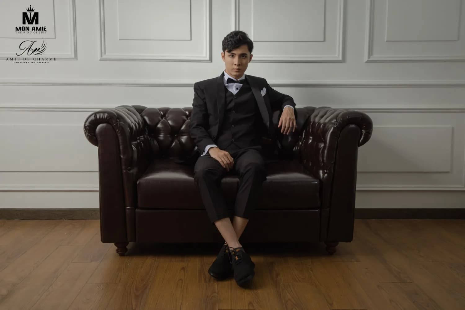 Tuxedo ba mảnh ngồi ghế sofa da nâu, tường phào chỉ trắng