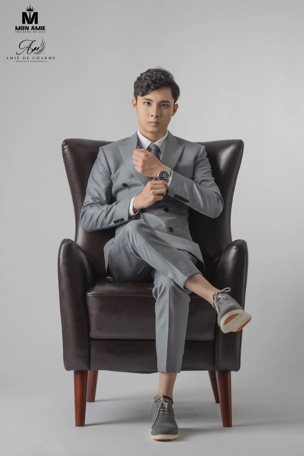 Suit xám hai hàng khuy, vắt chân trên ghế bành da nâu, chỉnh đồng hồ